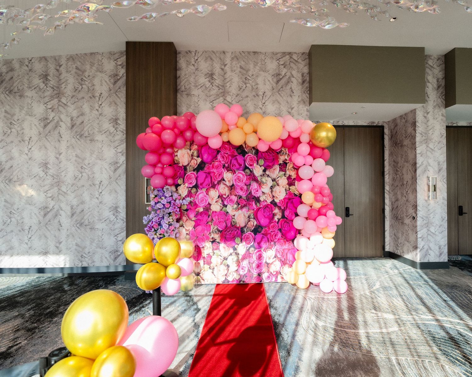 A colorful balloon arch installation in progress shows pink, yellow and red balloons being arranged against a concrete wall.