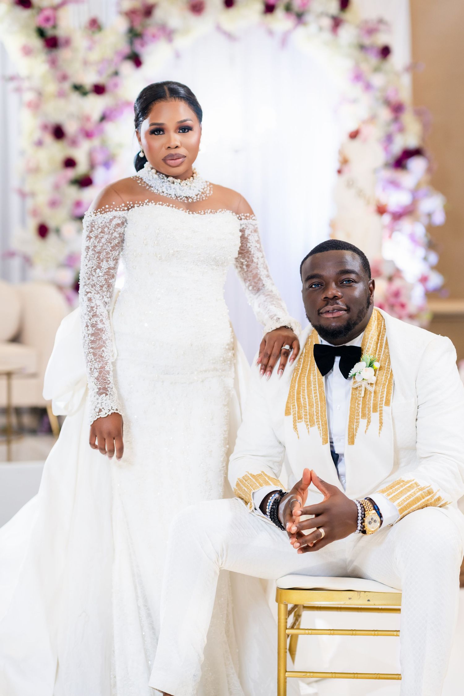 Wedding couple in matching white attire pose together against floral backdrop with gold chairs.