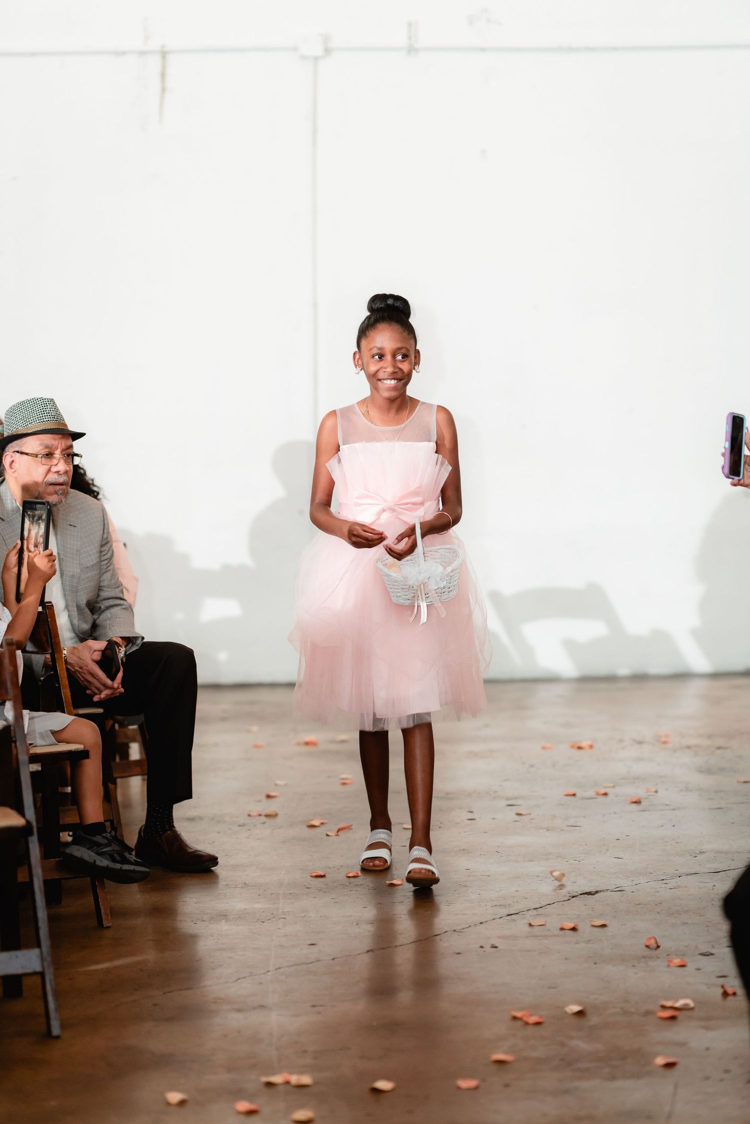 Wedding party members sit in chairs on a white floor scattered with rose petals.