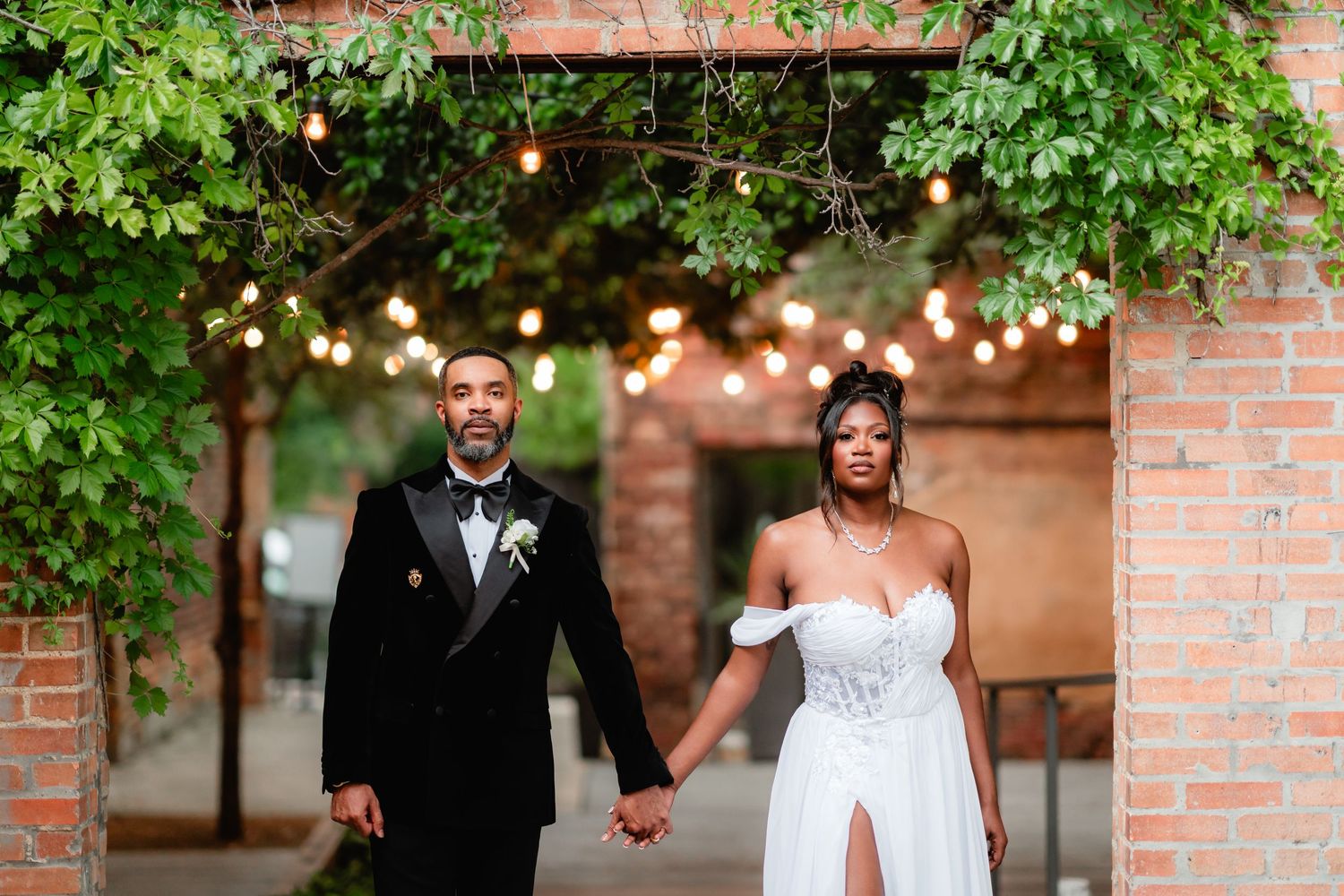 Newlyweds walk through a garden setting with string lights and greenery creating a romantic atmosphere.