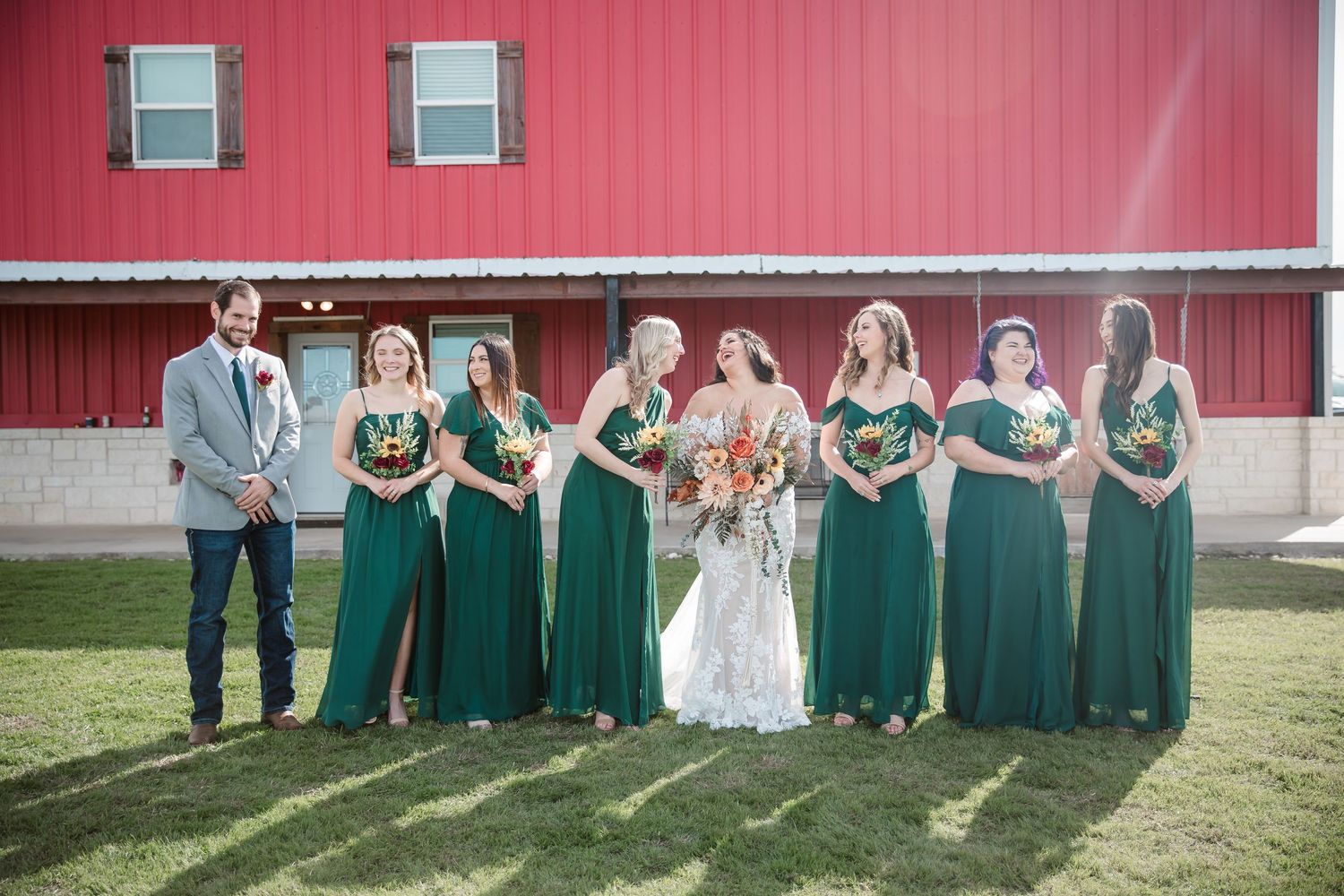 Bridesmaids in emerald green dresses pose against red barn wall.