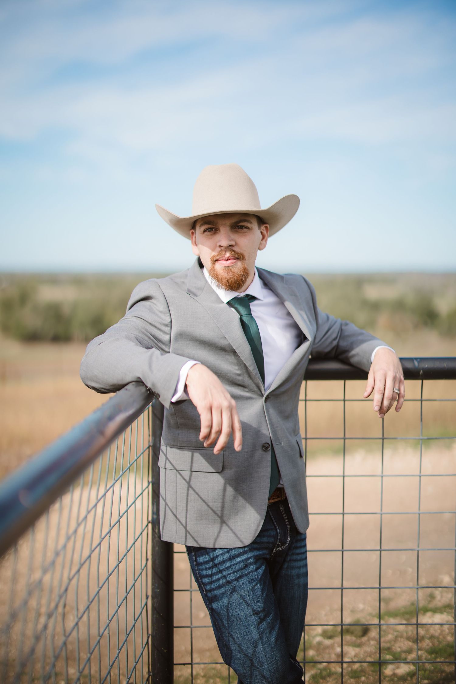 Cowboy in gray suit and hat leans against rustic fence in rural landscape.