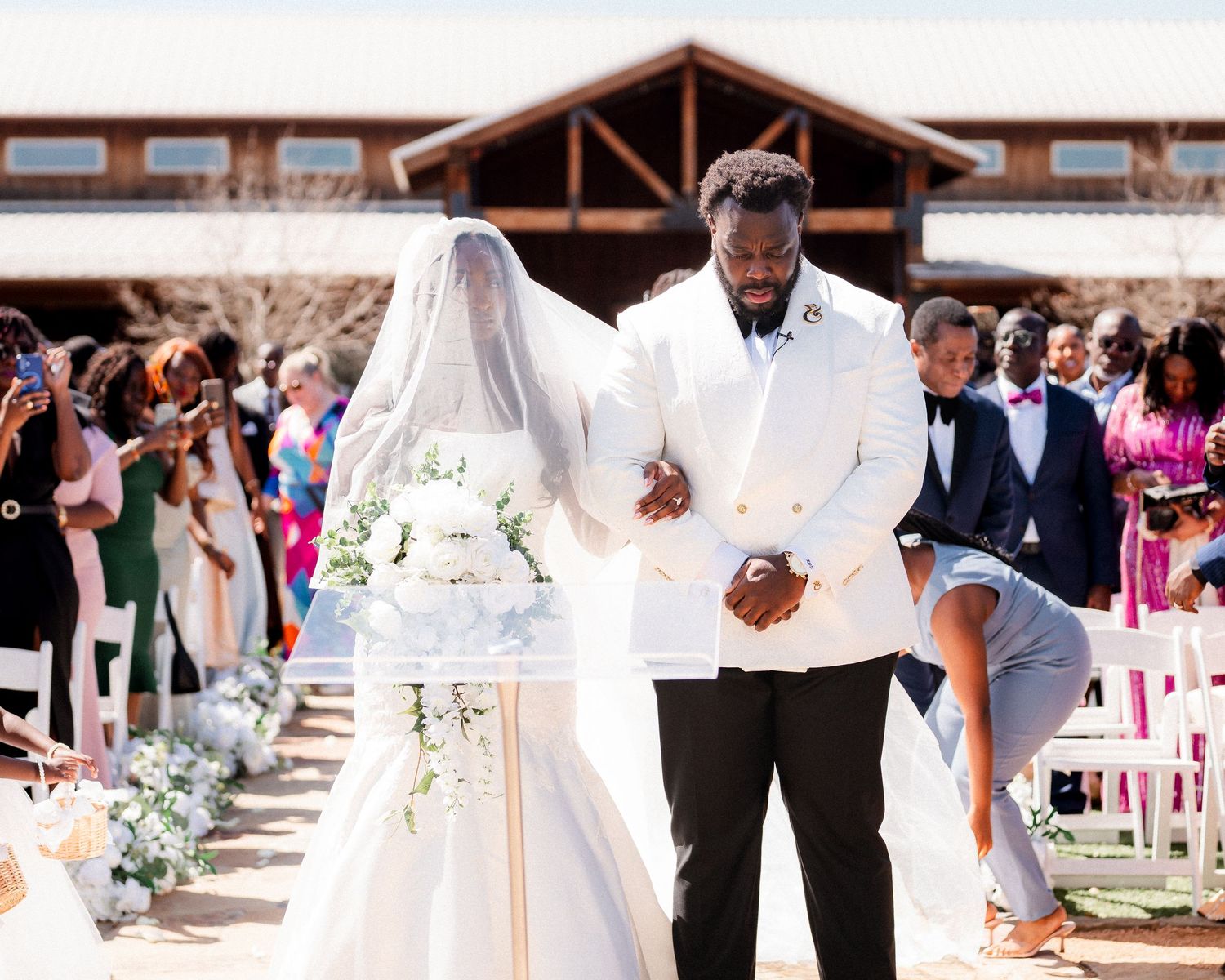 A wedding ceremony takes place outdoors with guests seated in white chairs and floral decorations along the aisle.