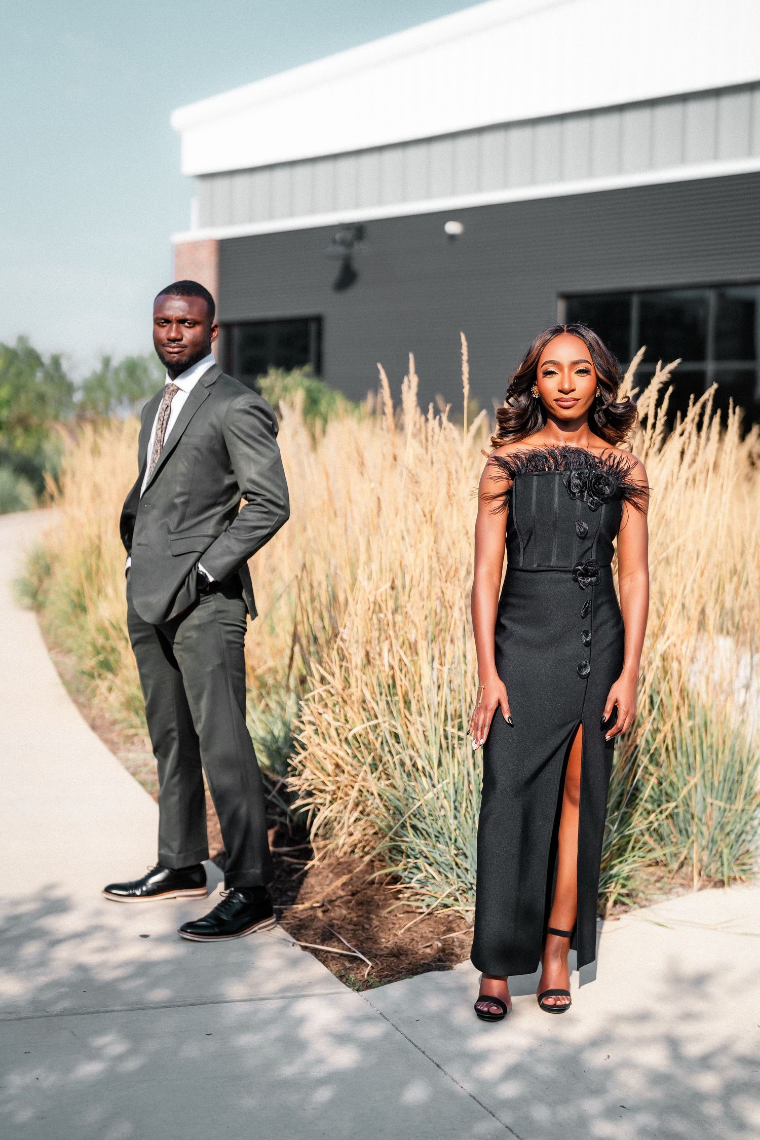 A fashionable couple poses separately in formal wear near ornamental grasses and modern architecture.