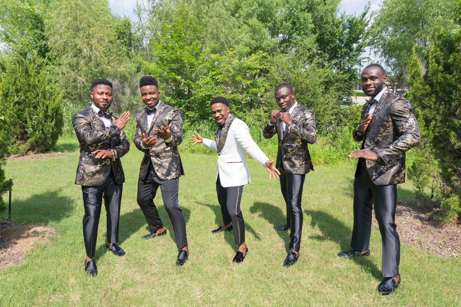 A group of people in formal attire jump and pose playfully outdoors in a green garden setting.
