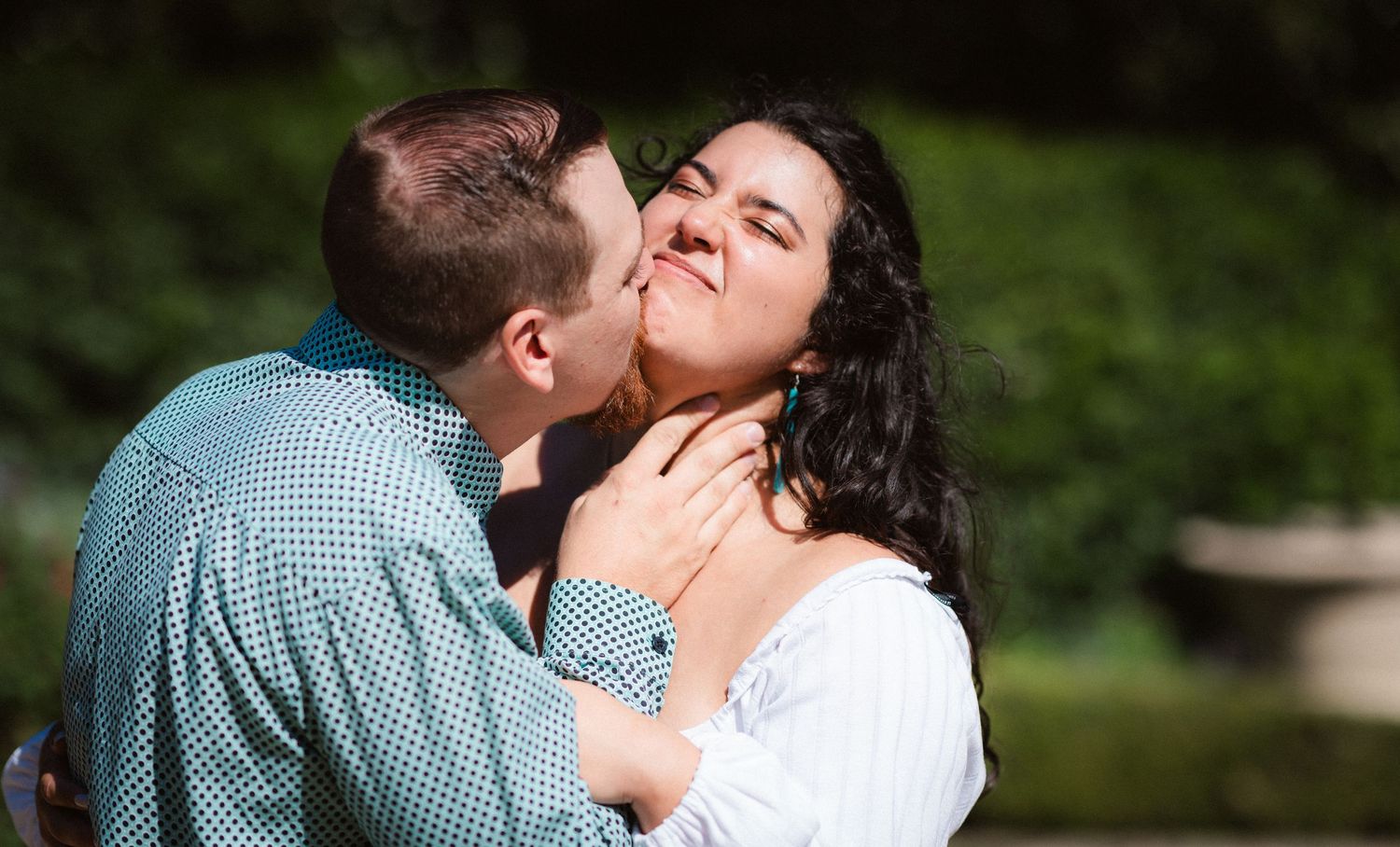A romantic couple shares tender moments in a lush green garden setting on a sunny day.