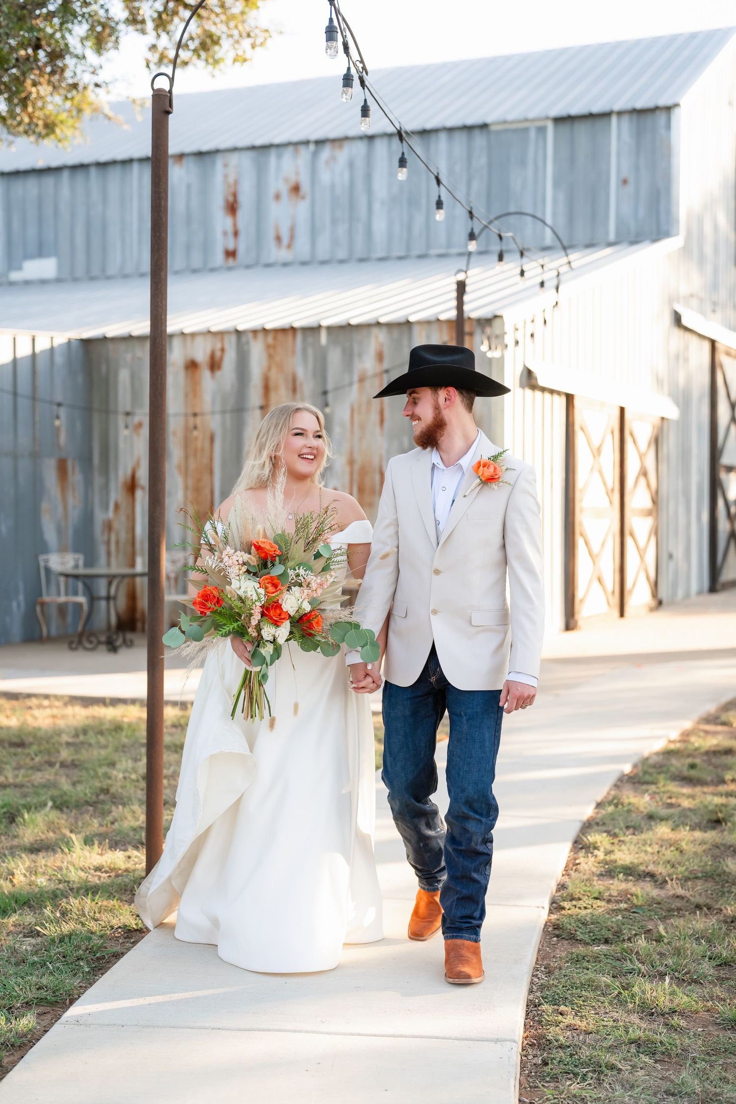 A rustic barn wedding scene with a couple in white attire walking along a metal building in warm sunlight.