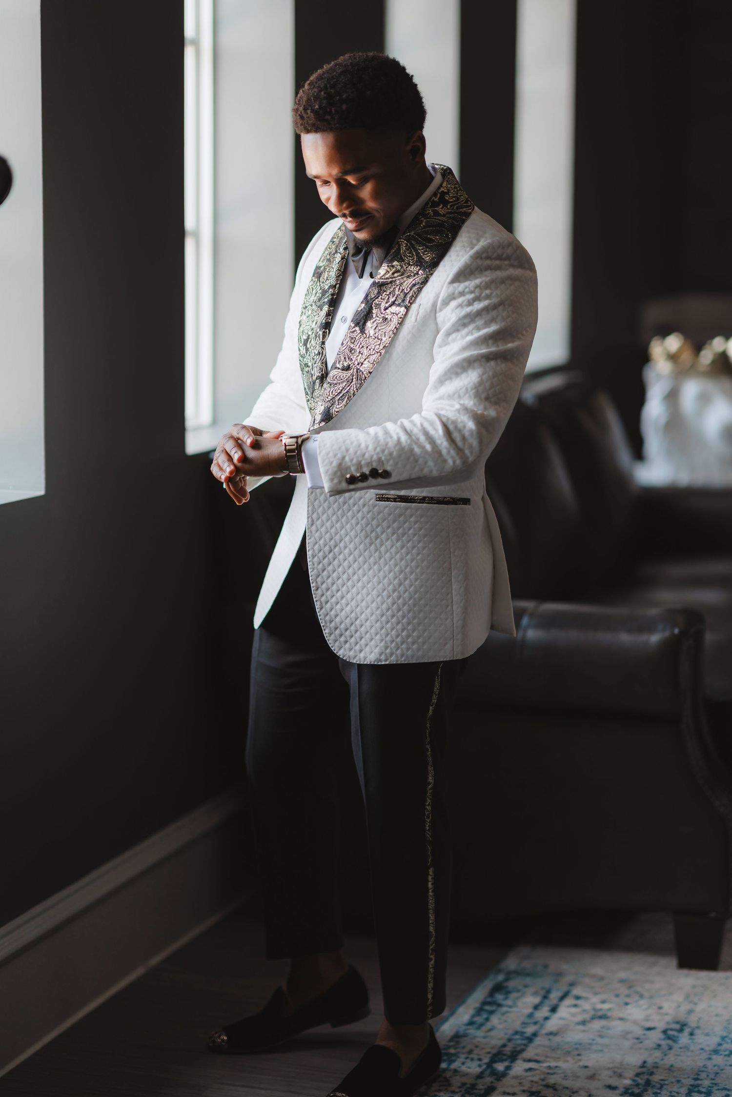 A person in a white textured blazer with floral lapel checks their watch by a window.