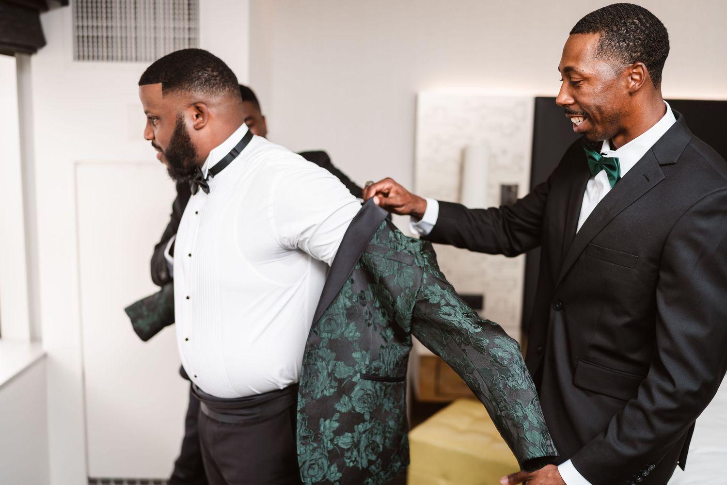 Two men in formal attire getting dressed, with one helping the other put on a green patterned jacket.