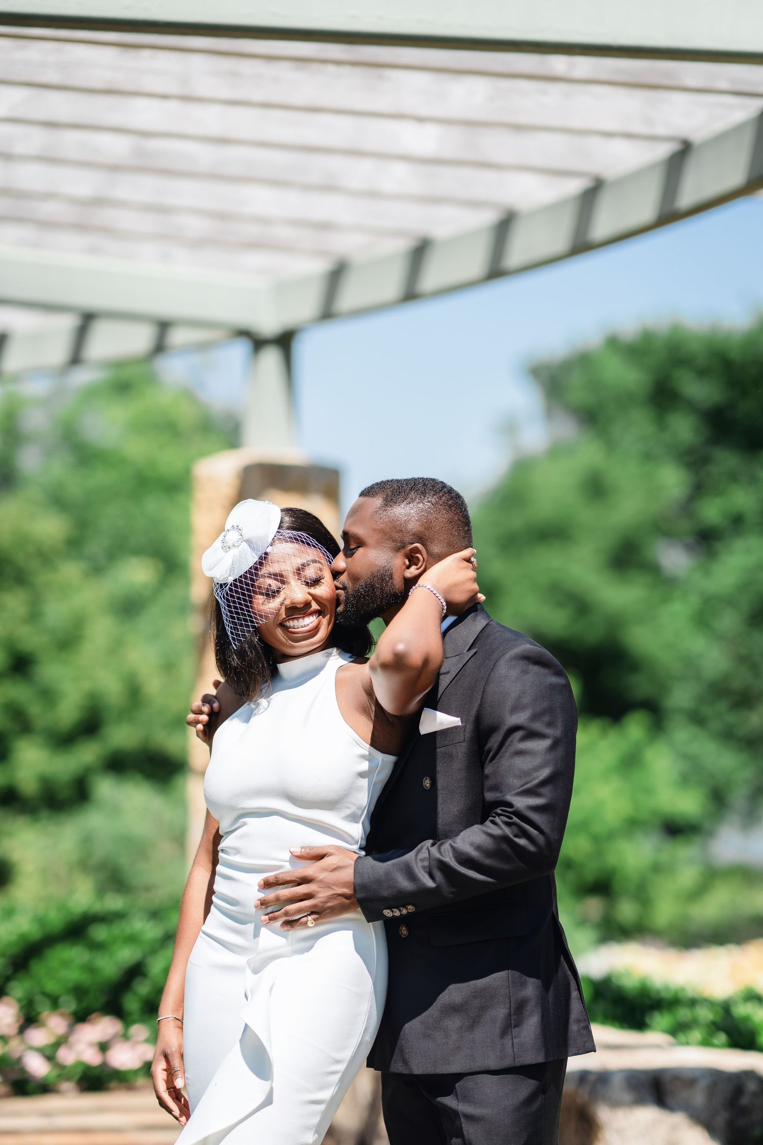 A couple shares a romantic moment during their wedding photoshoot under a white pergola with greenery in the background.