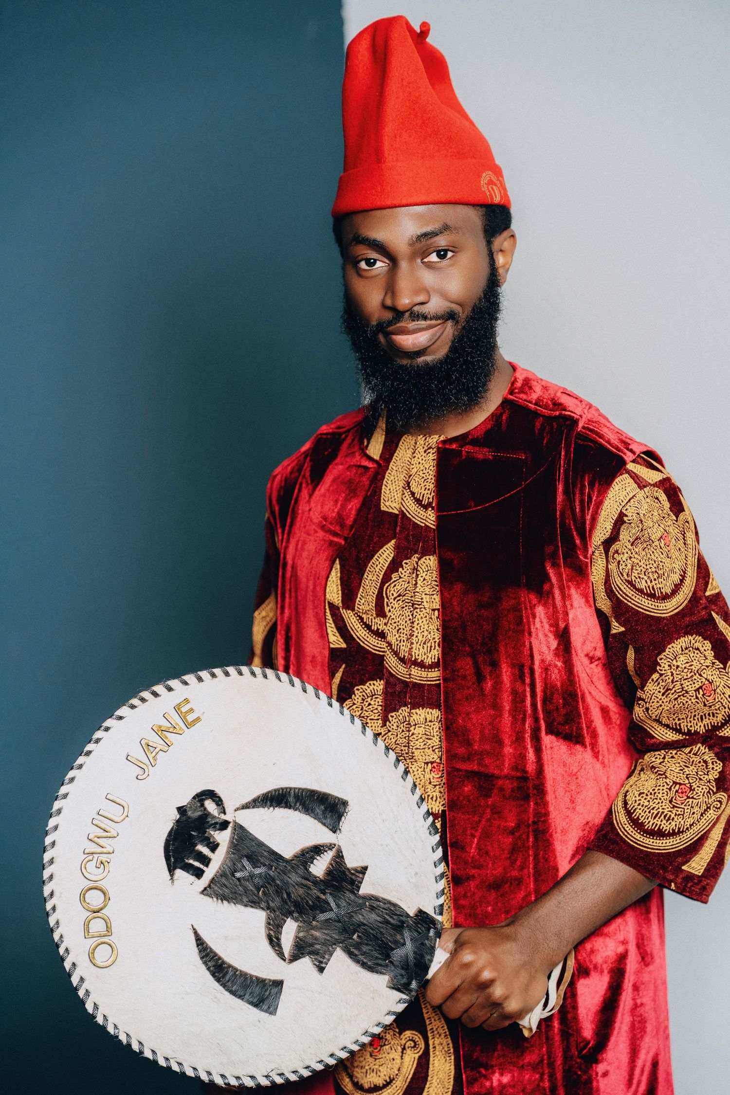 Traditional African groom in red velvet attire and cap displaying ceremonial fan against grey background.