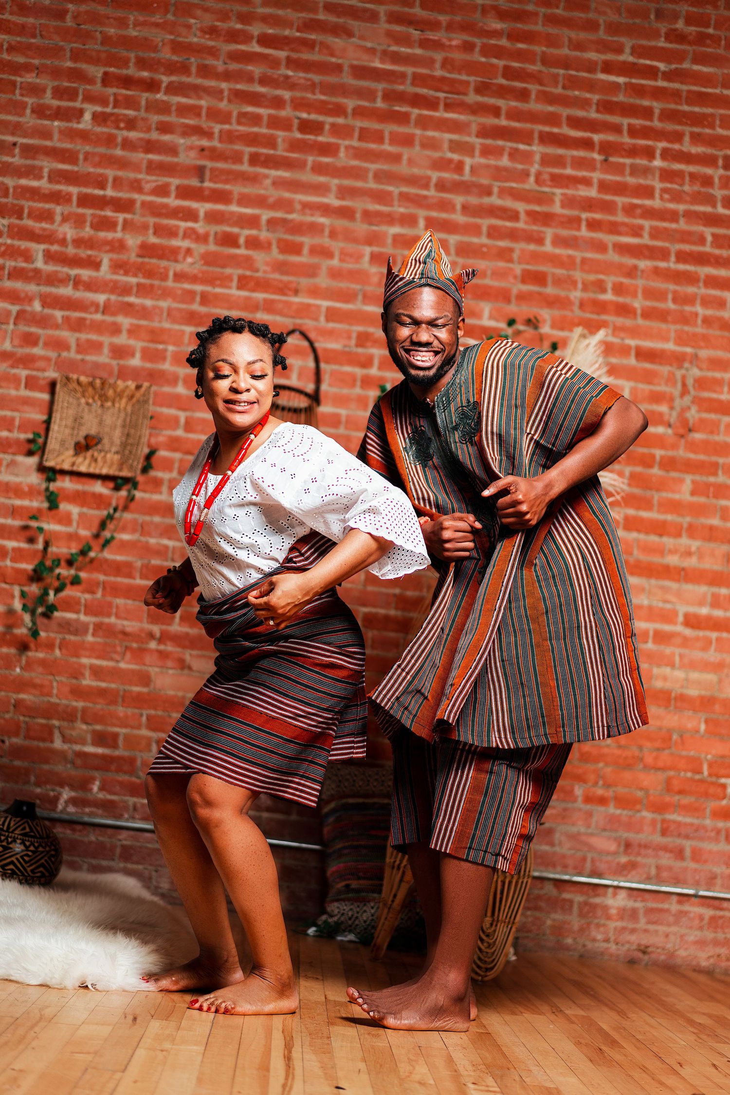 A couple in traditional African attire dance and pose playfully against a brick wall background.