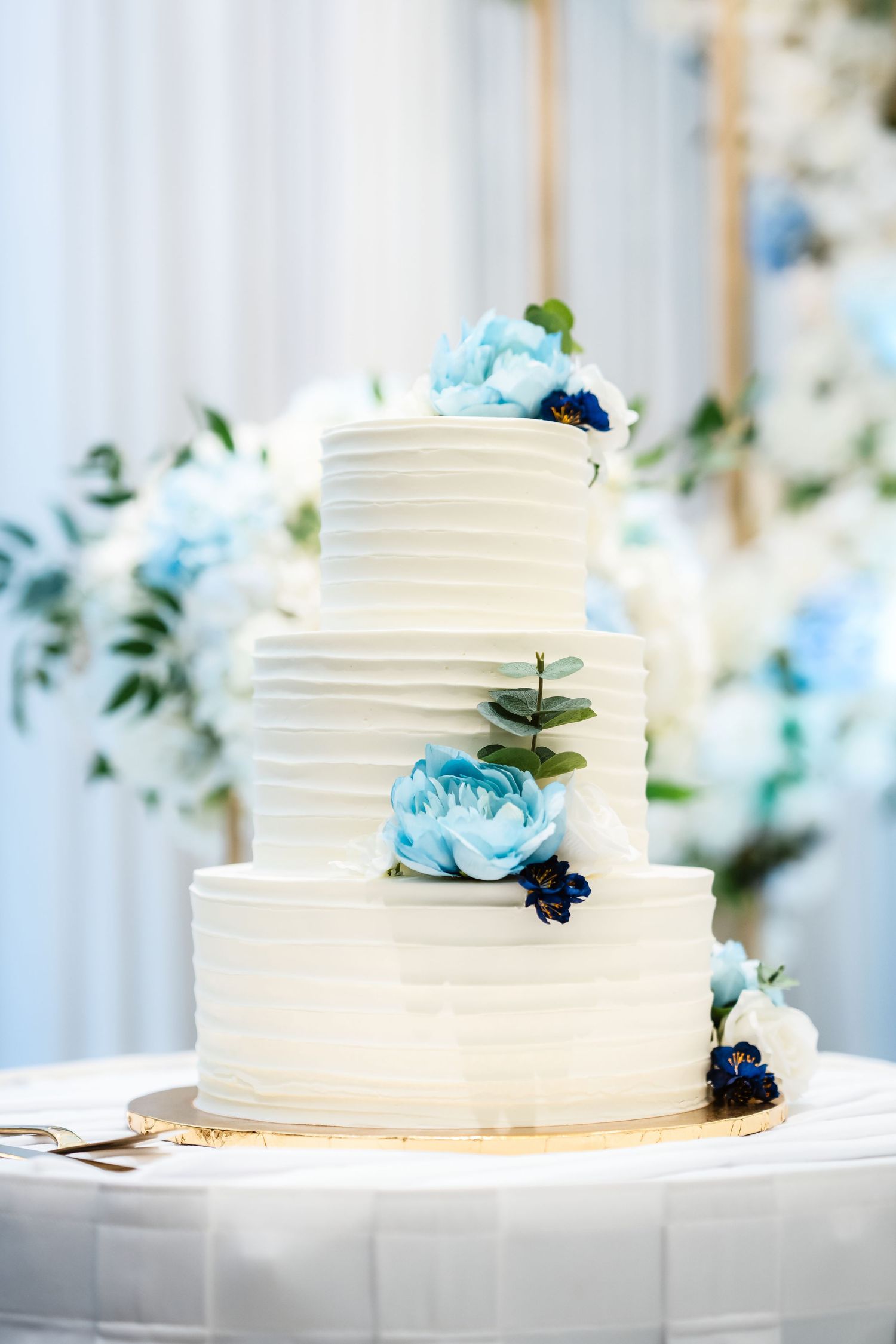 A white tiered wedding cake decorated with light blue flowers and greenery.