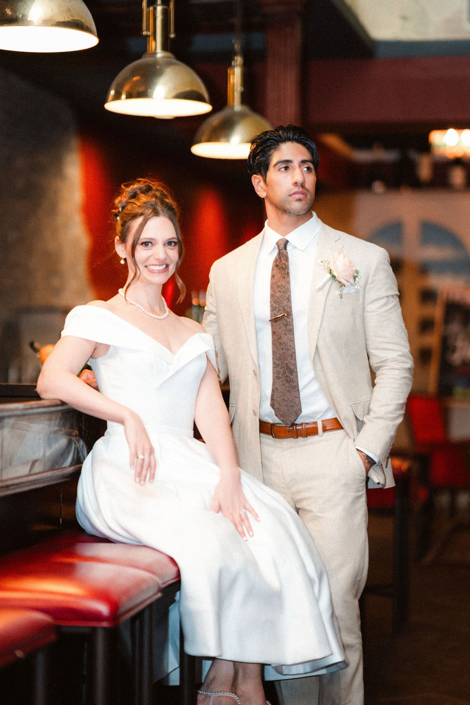 A couple in wedding attire poses at a bar with pendant lighting and red leather seating.