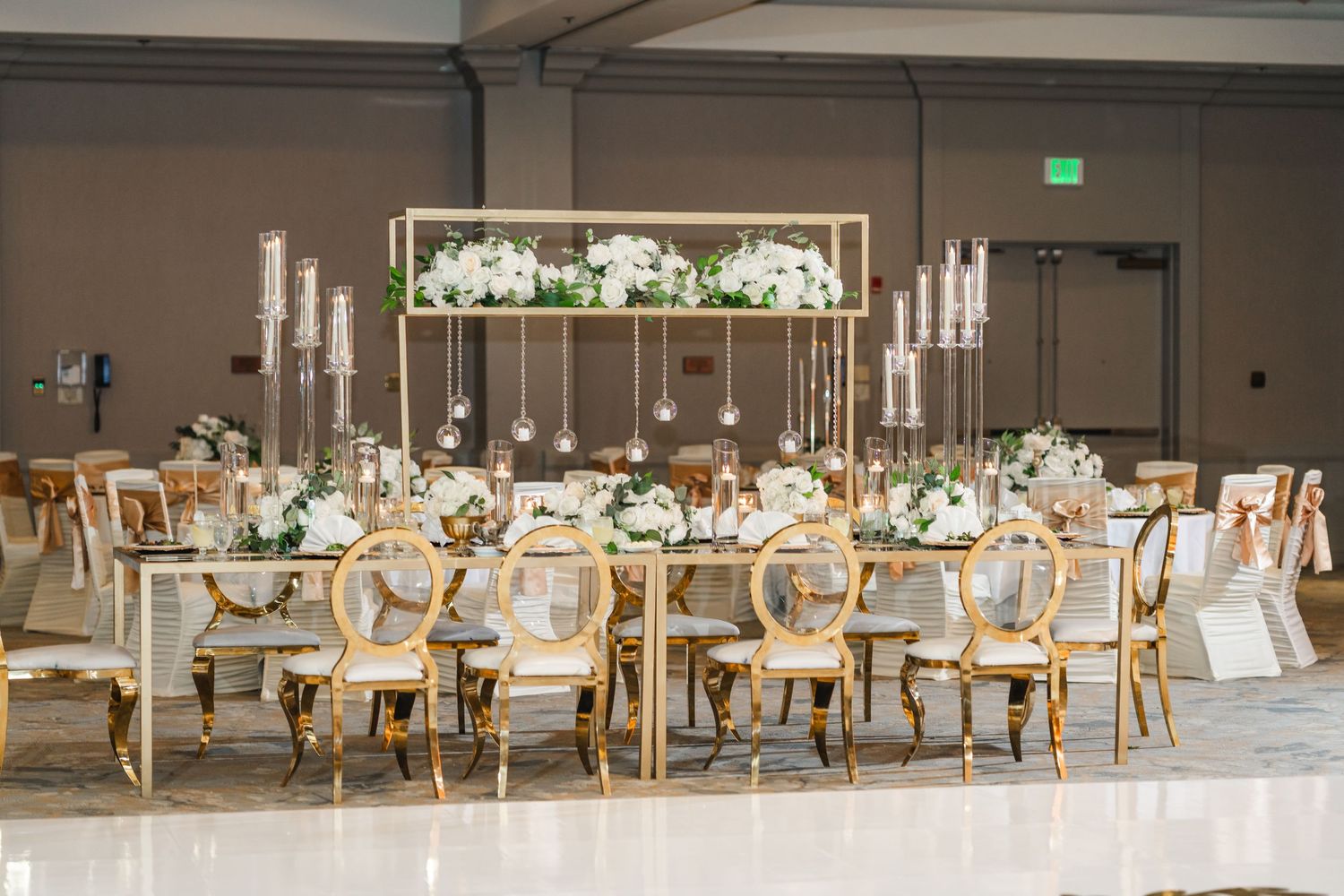 Elegant wedding reception setup with gold chairs and white floral centerpieces in a spacious ballroom.
