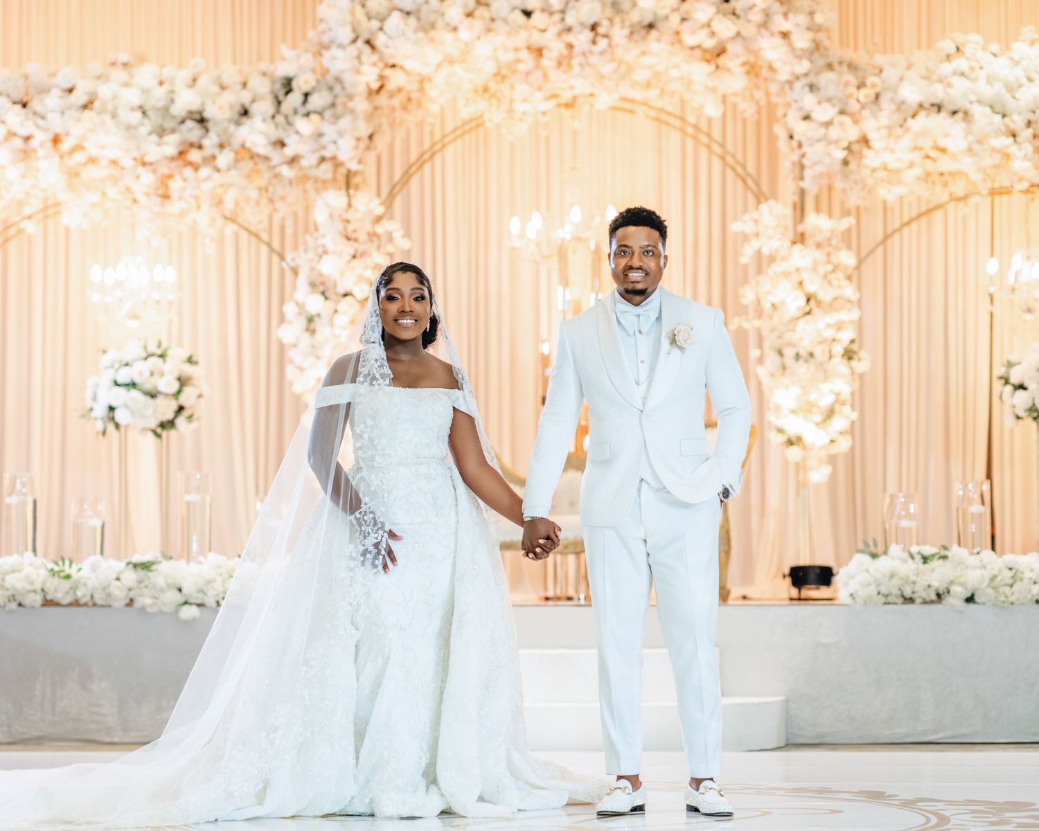 A wedding couple poses in elegant all-white attire against a floral backdrop in a romantic setting.