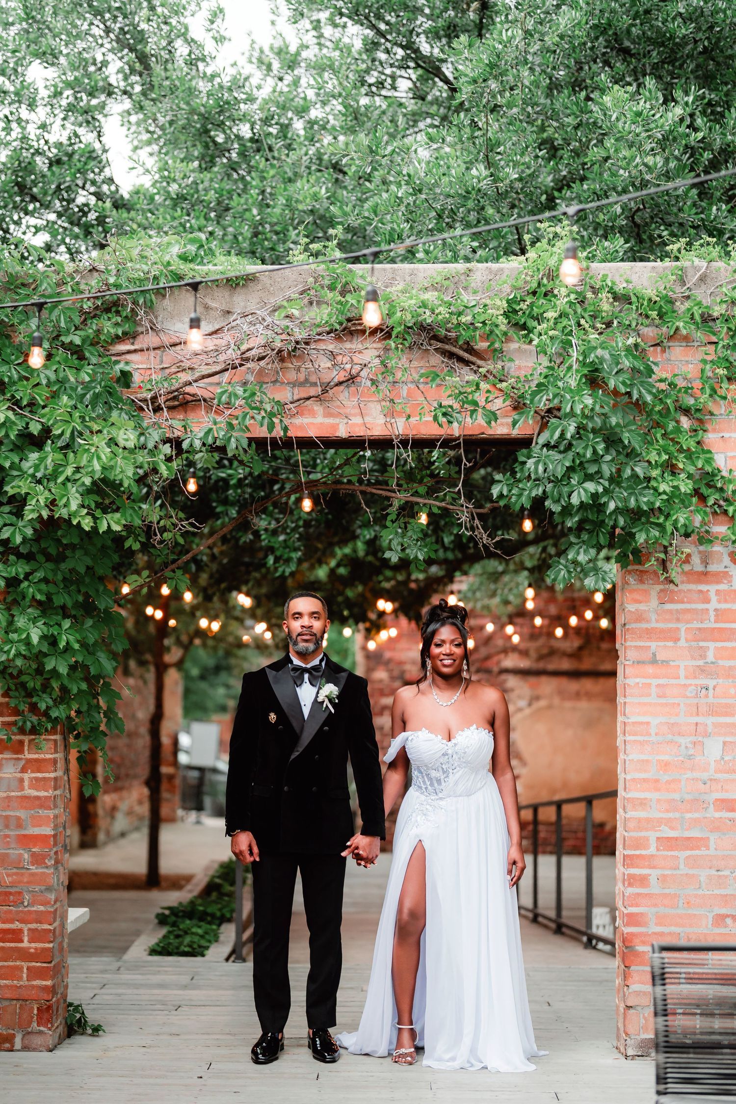 Wedding couple walking under string lights and greenery along a brick-lined outdoor walkway.