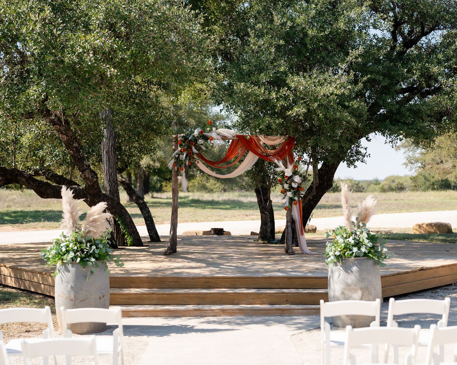 A serene outdoor wedding venue with white chairs arranged under shady trees.