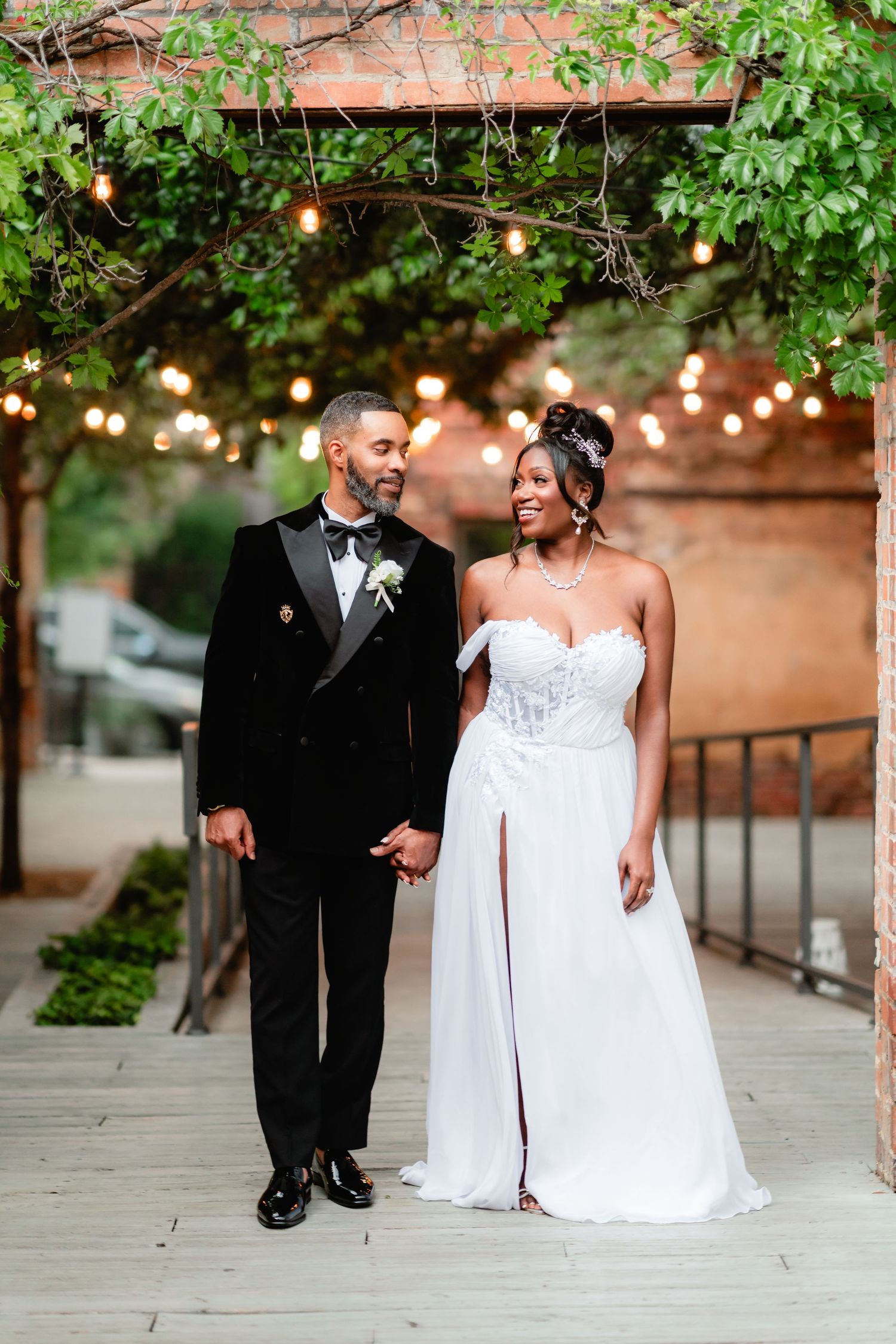 Elegant couple walks together on a brick-lined path under twinkling string lights.