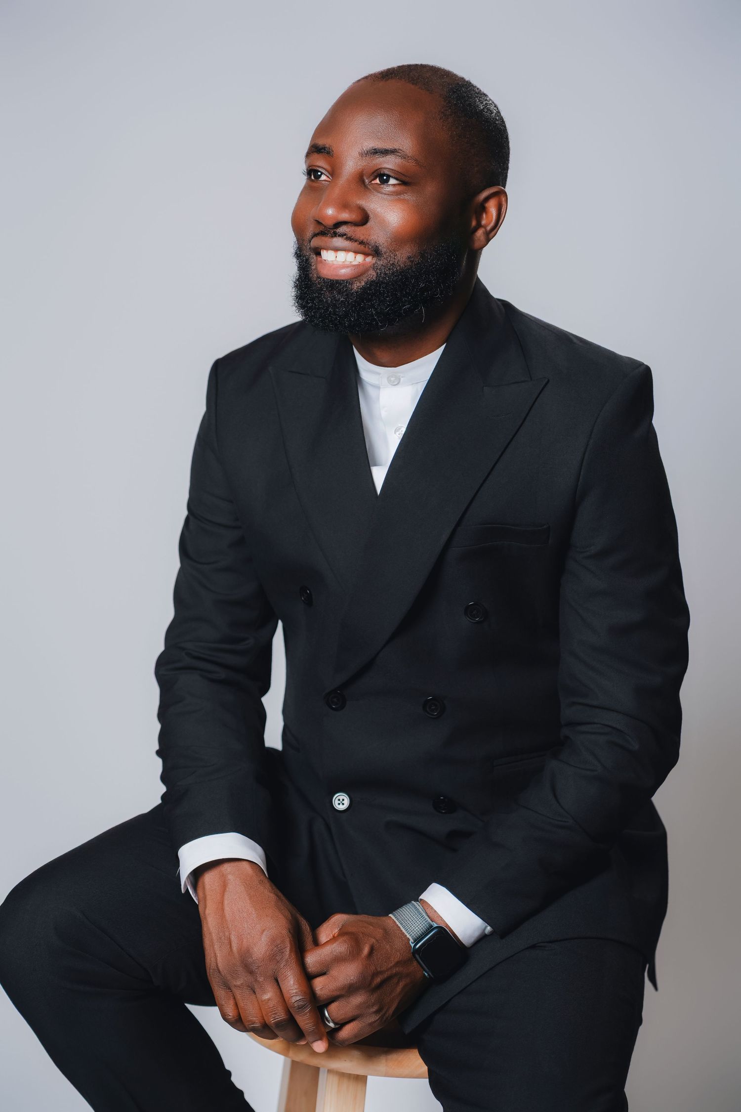 Professional portrait of a man in a black suit and white shirt sitting casually against a gray background.