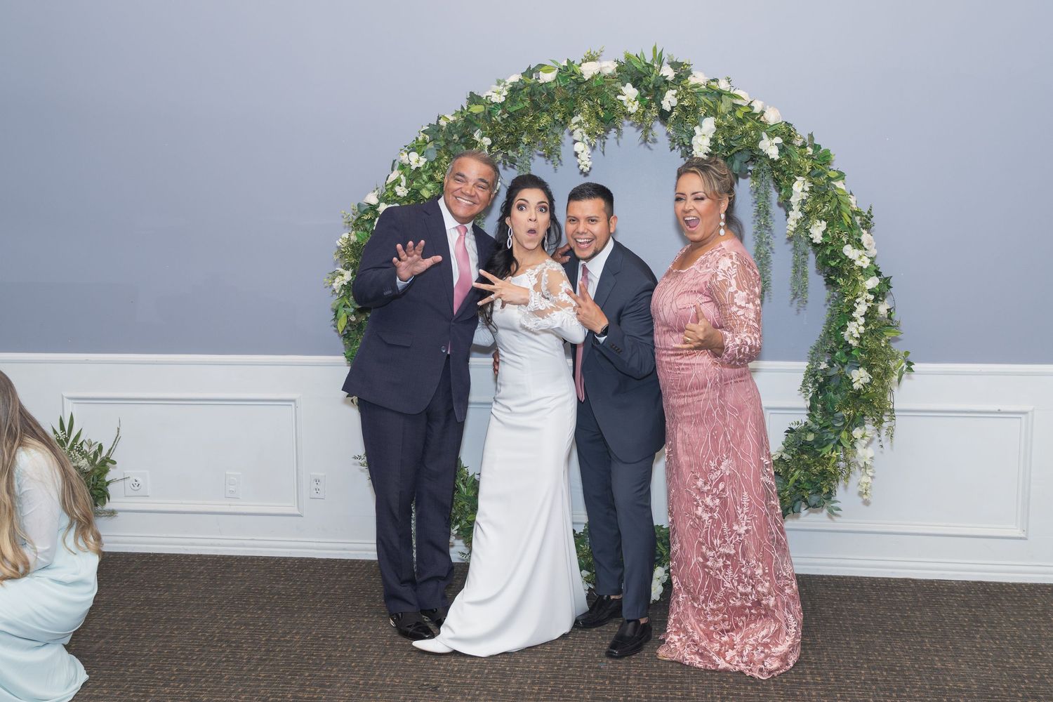 Wedding ceremony setup with floral arch and white chairs against grey wall.