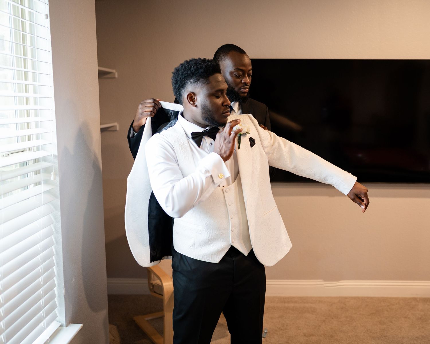 Groomsmen in black tuxedos and bowties adjusting their outfits in a sunlit room.