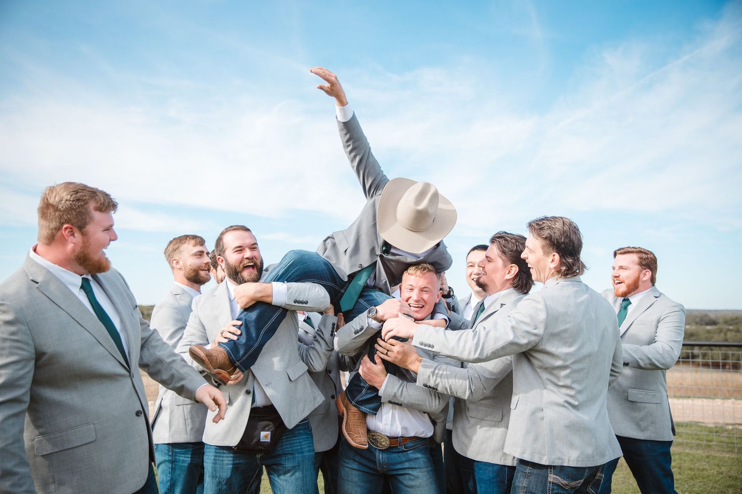 Groomsmen in light gray suits celebrating and jumping outdoors against a bright blue sky.