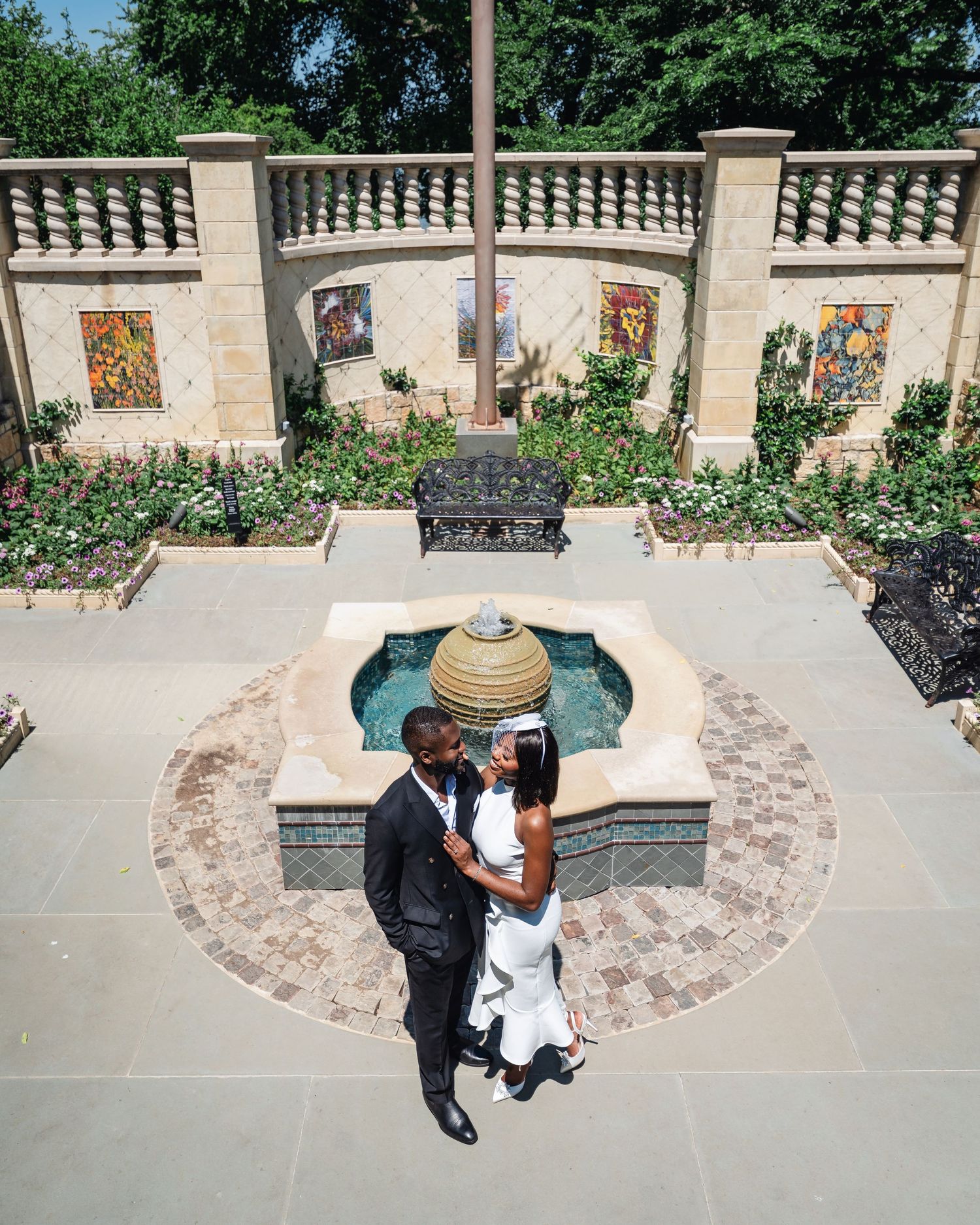 A couple stands in a garden courtyard with ornate wall decorations and a circular fountain feature.