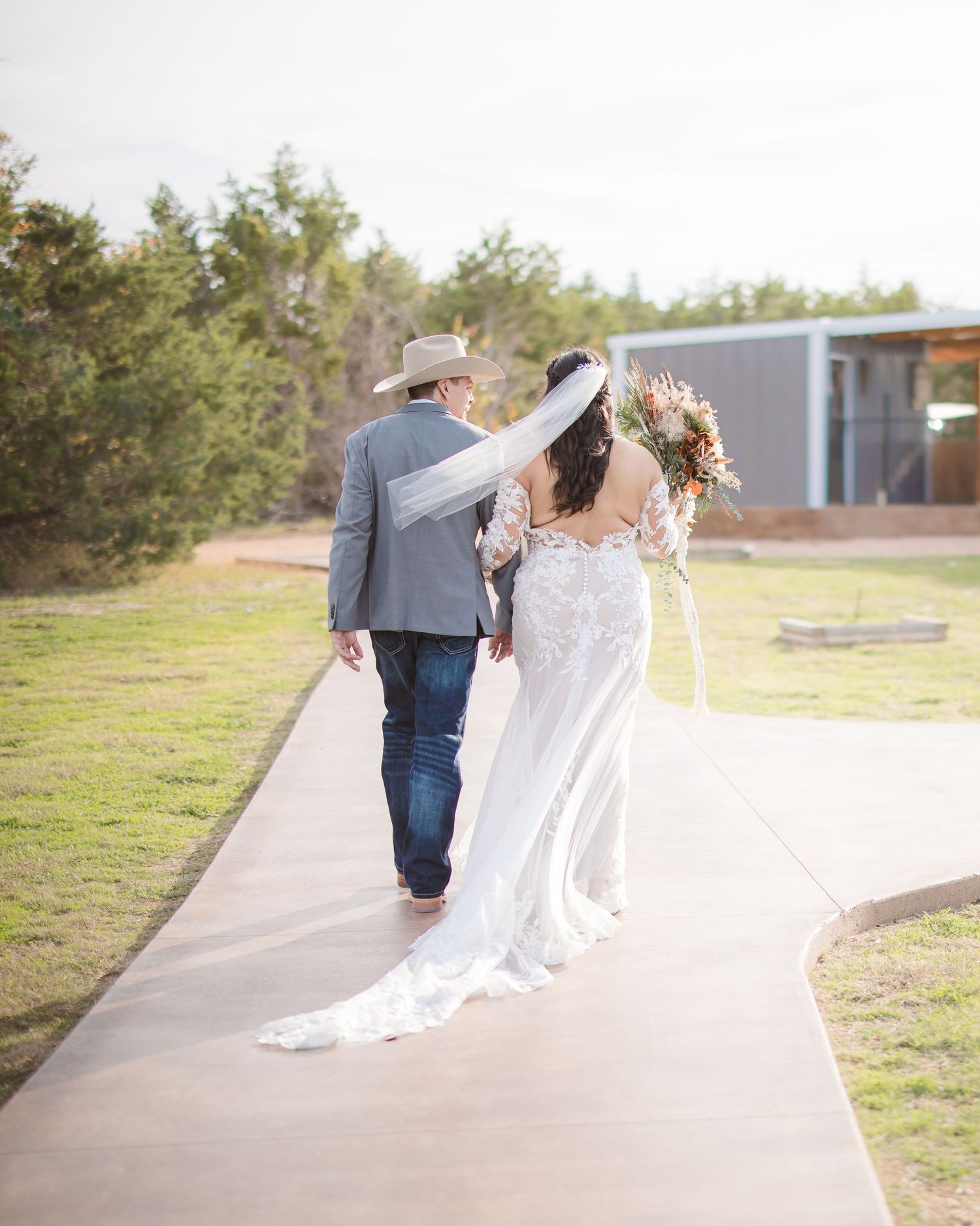 A couple walking down a concrete path outdoors, with the bride wearing a flowing white lace gown with a long train.