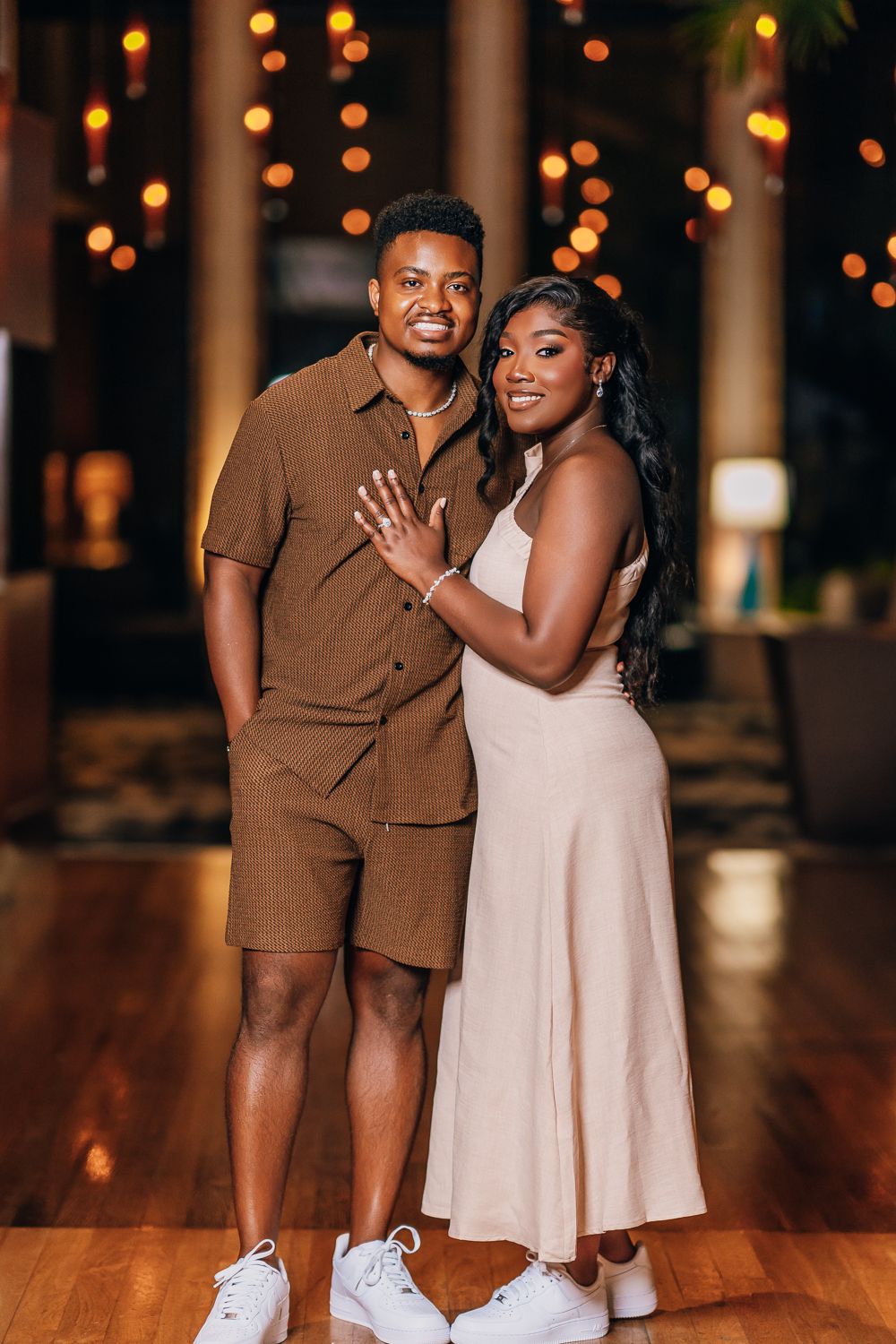 A couple in brown and white outfits poses playfully together under decorative string lights.