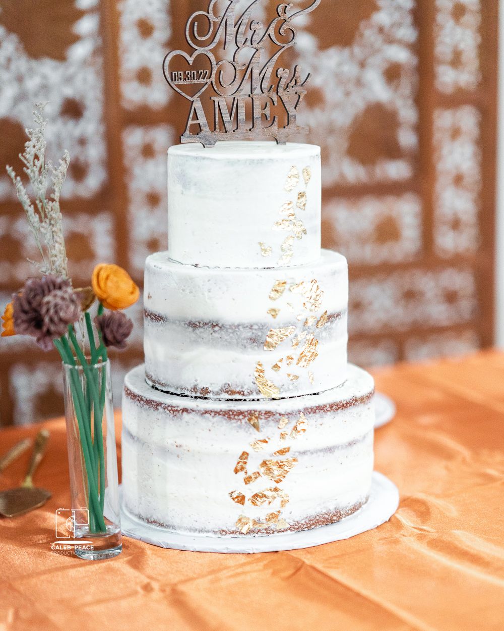 Three-tiered white wedding cake with delicate floral decorations displayed on an orange tablecloth.