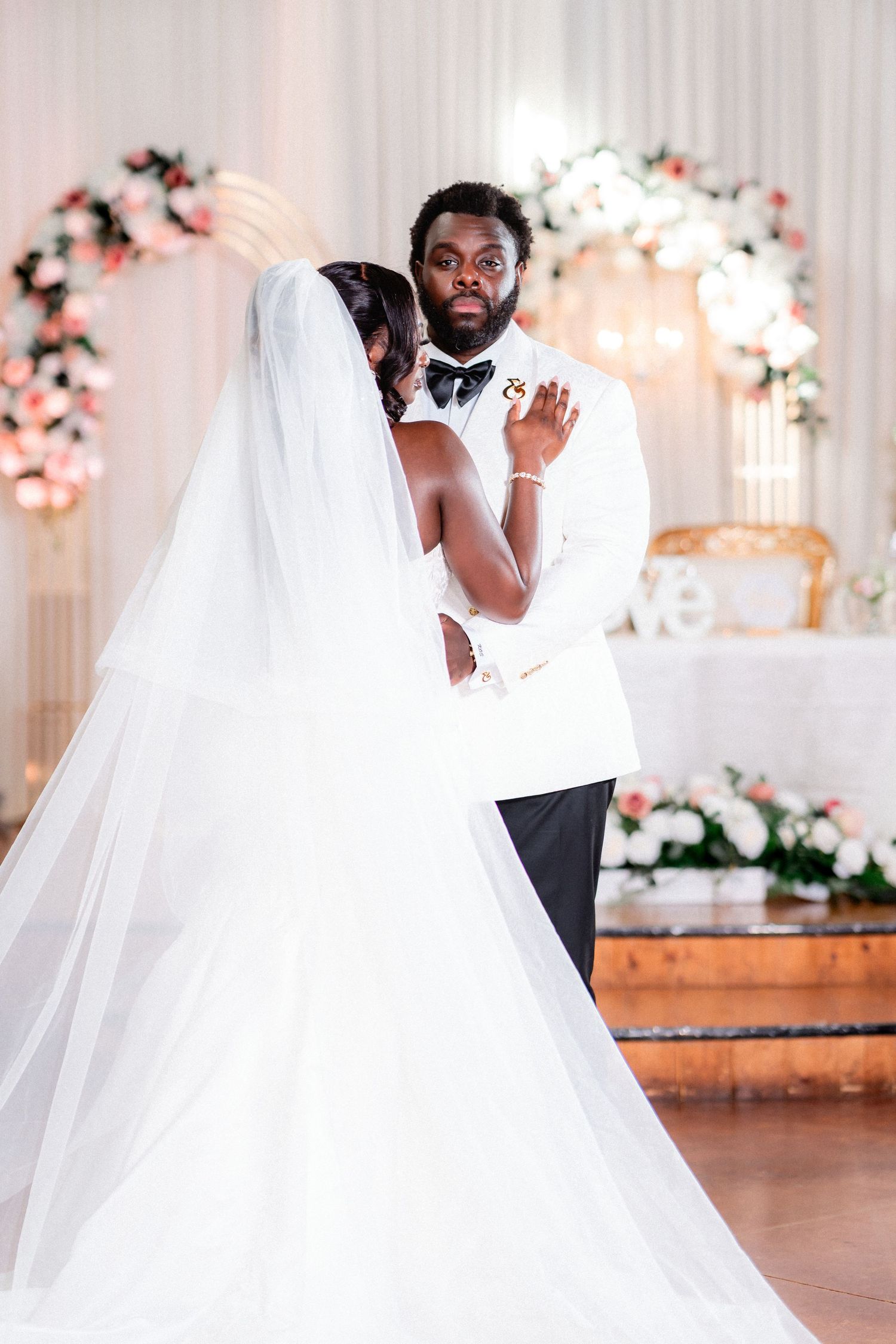 A romantic wedding moment captured in a white-decorated venue with floral arrangements and soft lighting.