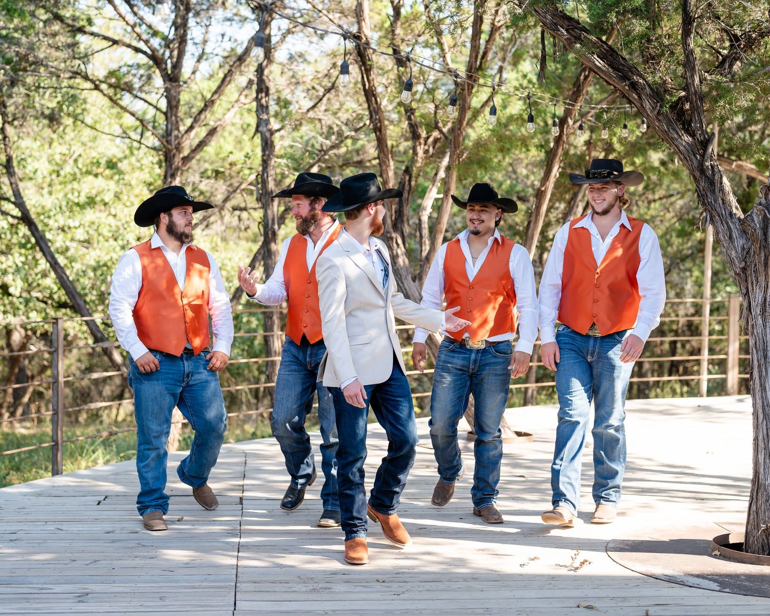 Groomsmen in orange vests and cowboy hats line up for a fun photo on a wooden deck.