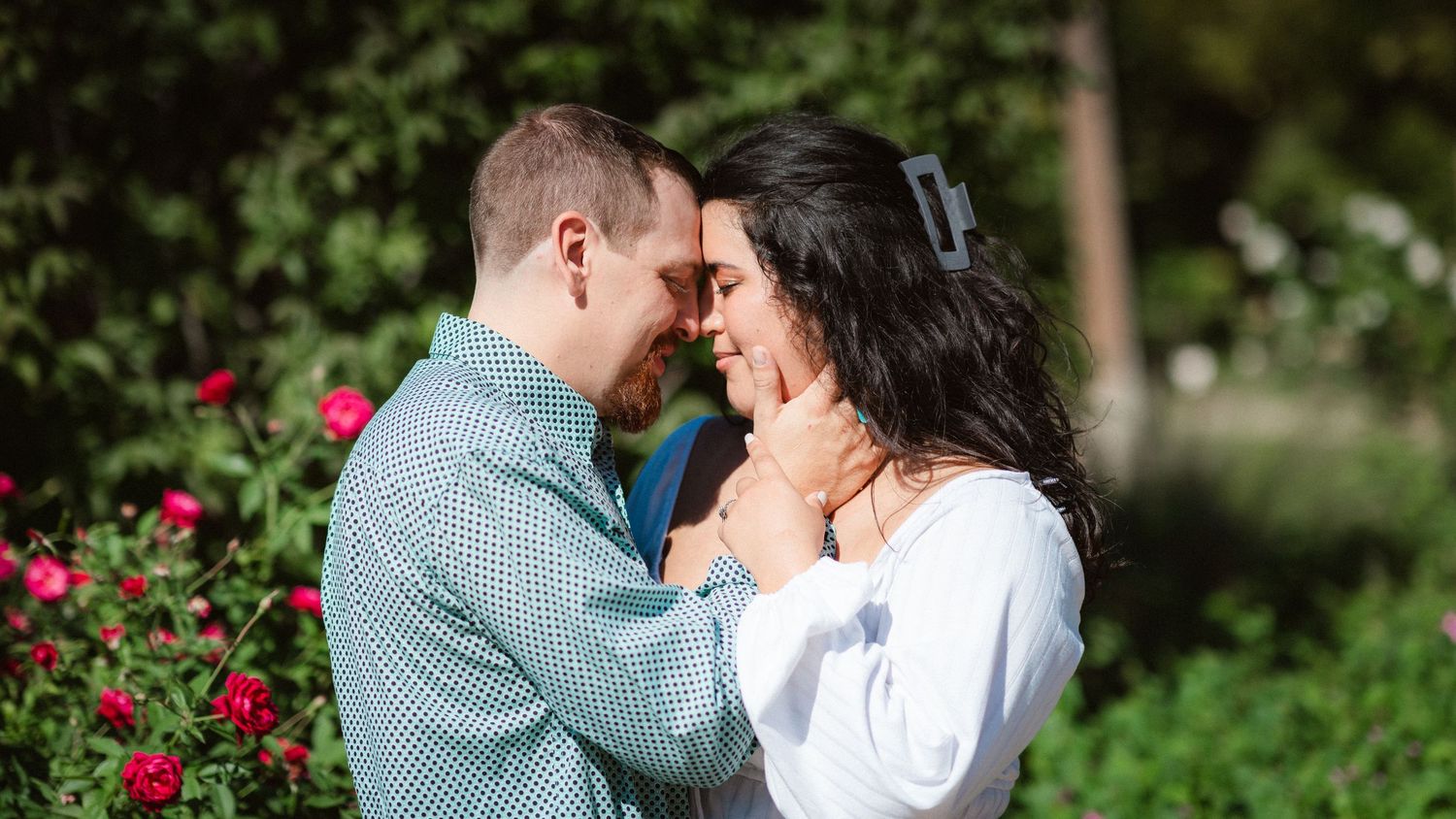 A couple shares intimate moments in a garden setting with vibrant red roses and lush greenery in the background.
