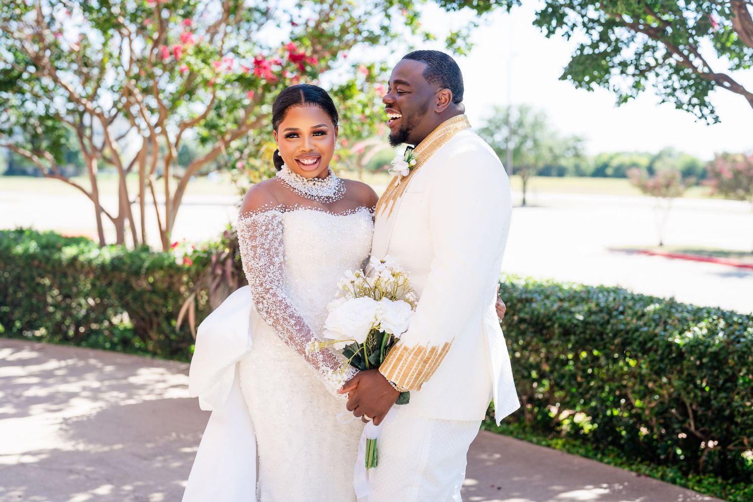A couple in elegant white wedding attire embrace outdoors on a sunny day with manicured greenery in the background.