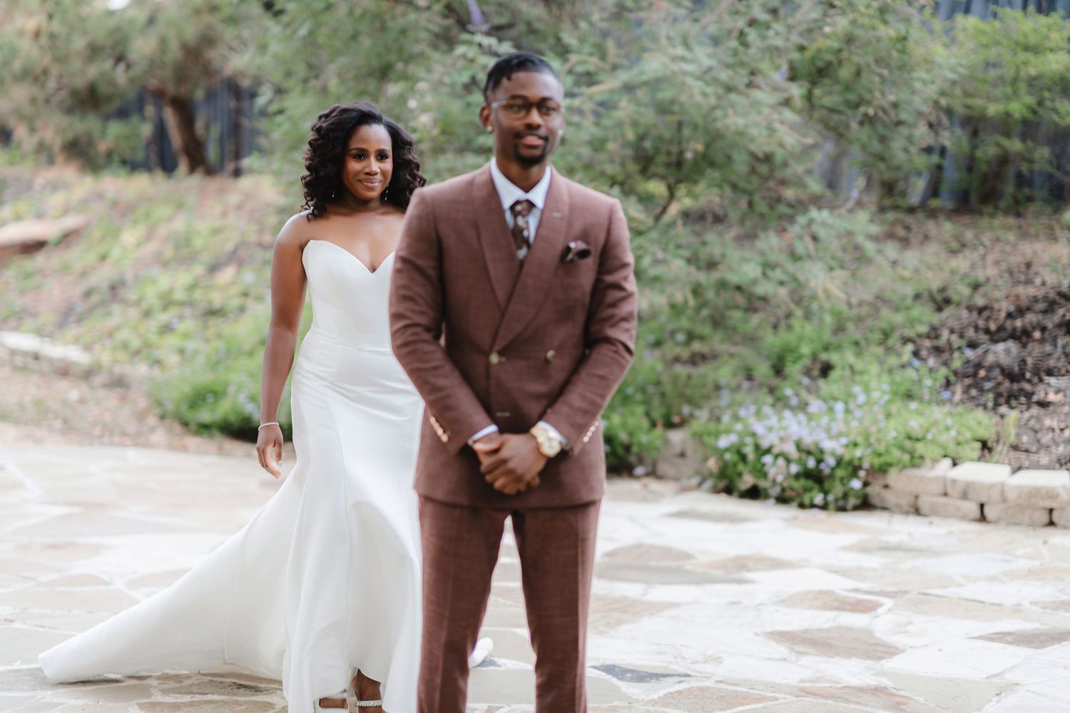 Newlyweds walk together on a garden path, with the bride in white and groom in brown.