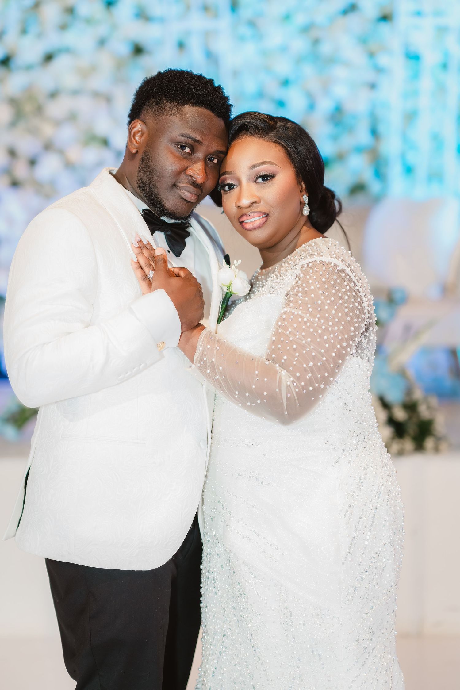 A romantic wedding portrait of a couple in white attire posing against a blue floral backdrop.