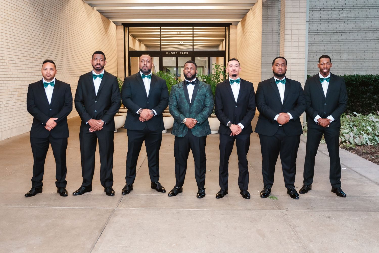 Groomsmen in black tuxedos seated in a row against a neutral wall.