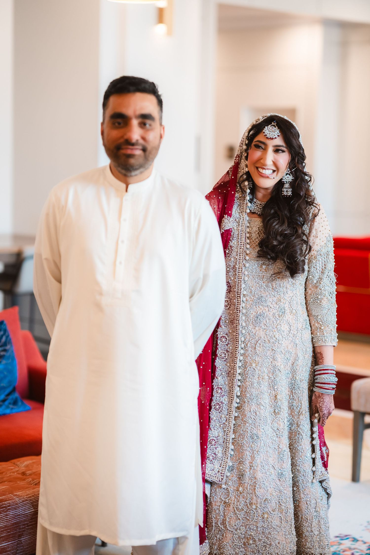 A couple in traditional South Asian wedding attire stands together in an indoor setting with red accents.