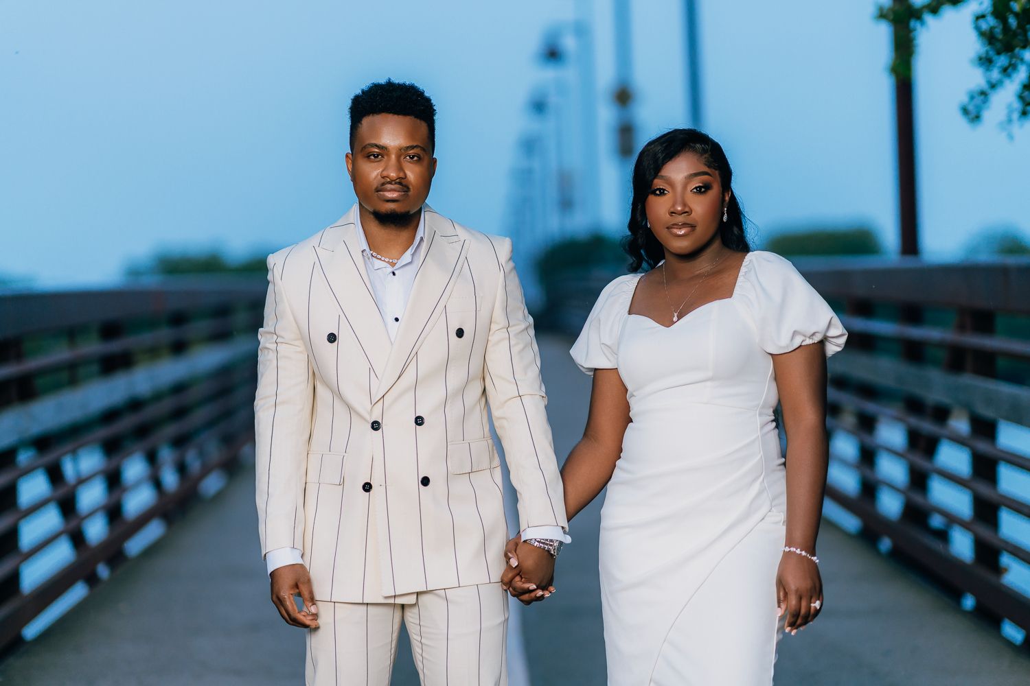 A couple in coordinating white and cream outfits pose together on a wooden boardwalk near water.