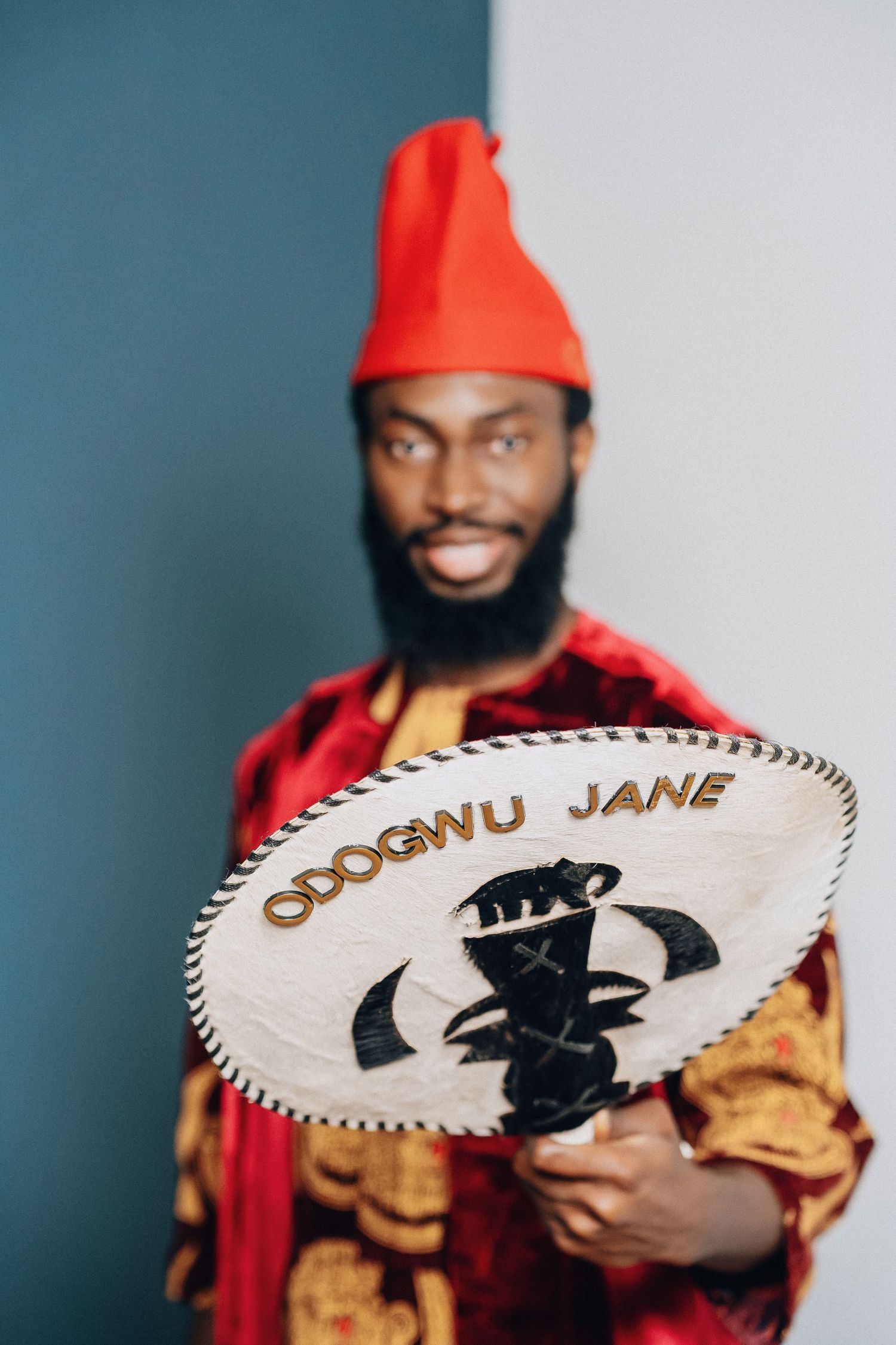 Person in red cap and velvet outfit holding decorative fan with traditional African designs.