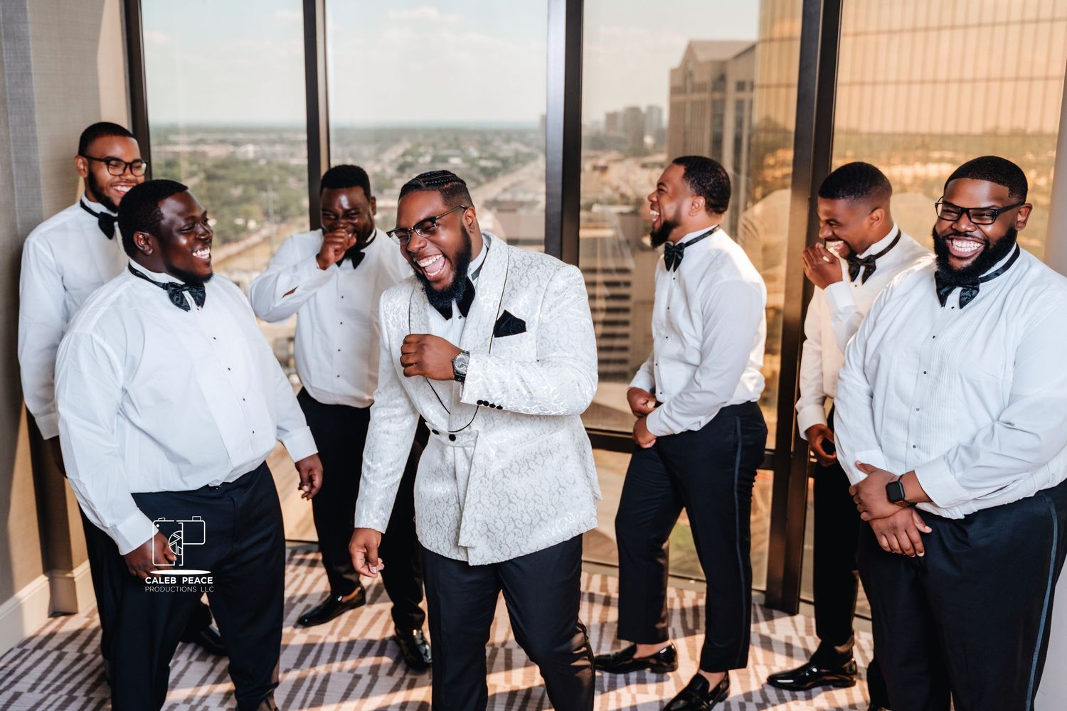 A group of groomsmen in white jackets share a laugh together on a high-rise balcony.
