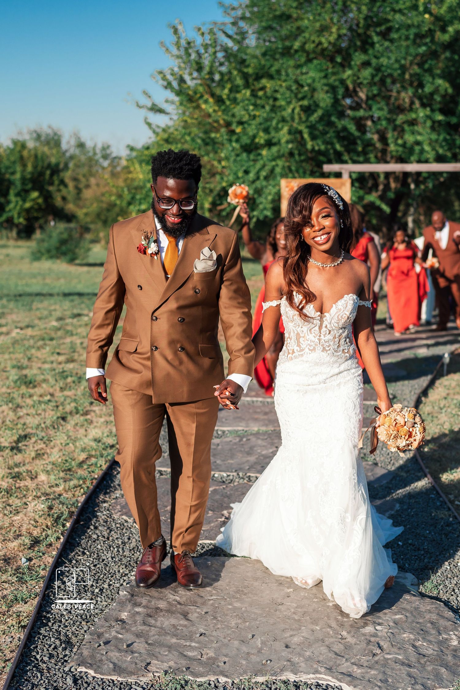 A couple walks together outdoors, with the bride in a white dress and groom in a brown suit.