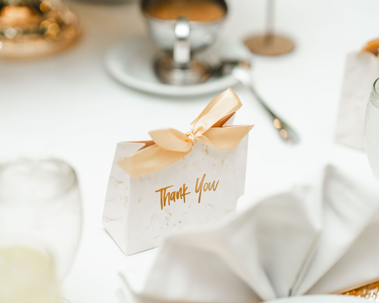Elegant white favor box with gold 'Thank You' text and peach ribbon on a table setting.