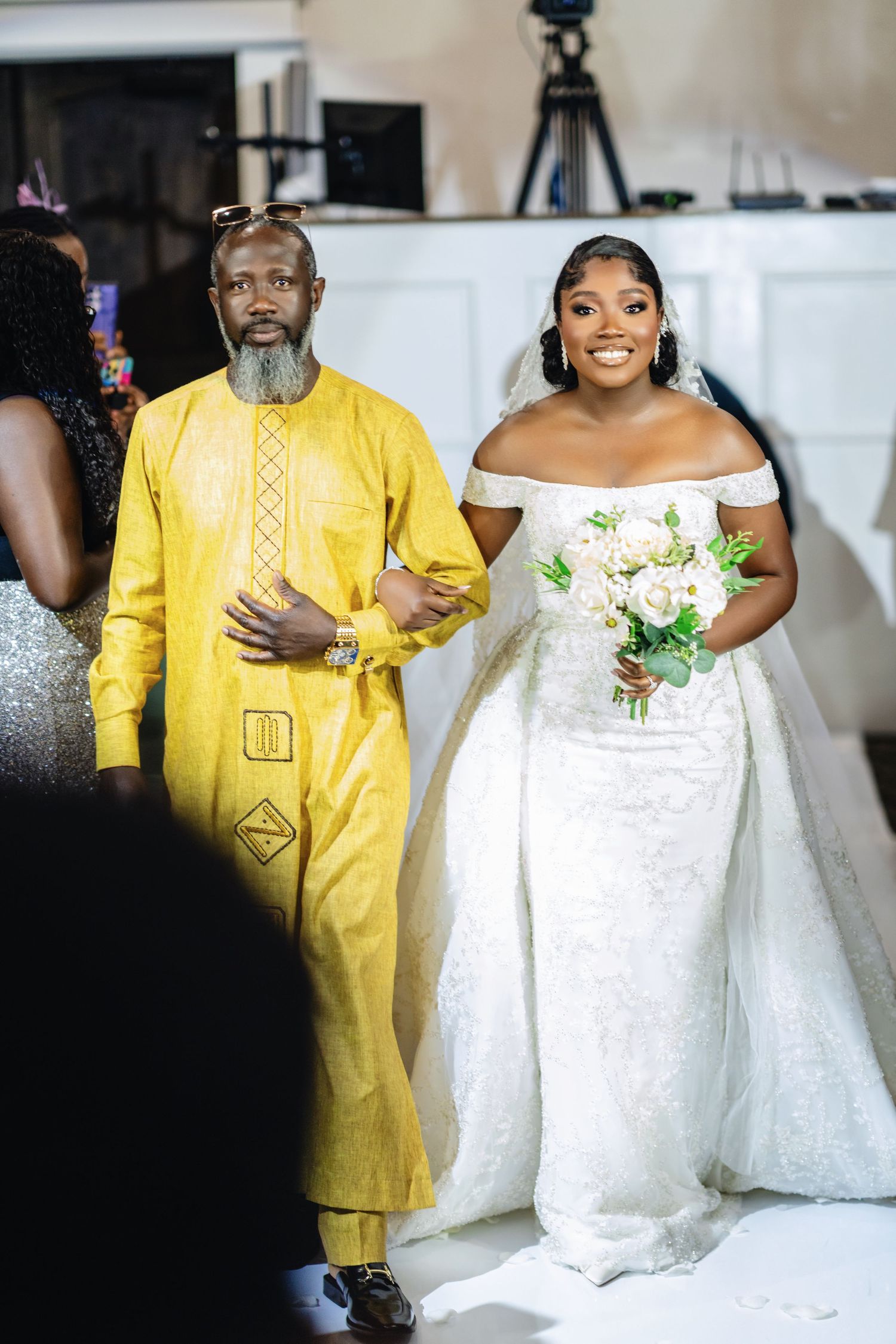 A bride in an off-shoulder gown walks down the aisle with an escort wearing a vibrant yellow traditional outfit.