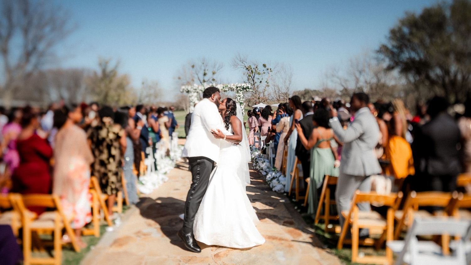 Outdoor wedding ceremony with guests seated on wooden benches under clear blue sky.