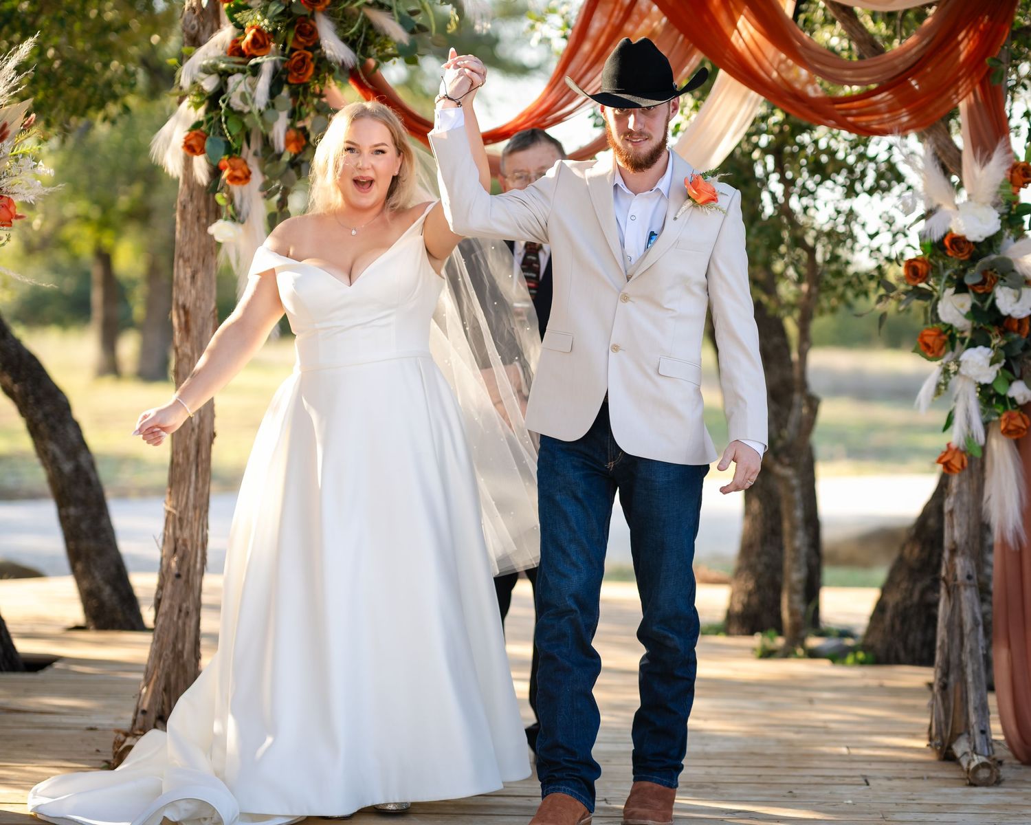 Wedding couple walking through tree-lined path with rustic barn in background.