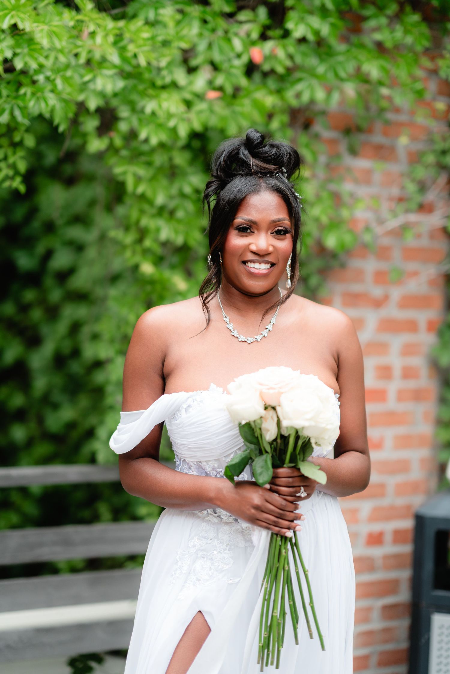 Beautiful bride in a white strapless gown poses with a bouquet against a green garden backdrop.