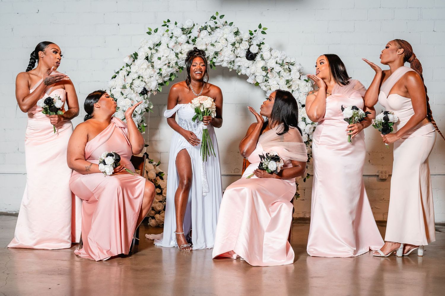 Bridesmaids in pink dresses pose playfully together against a white wall.