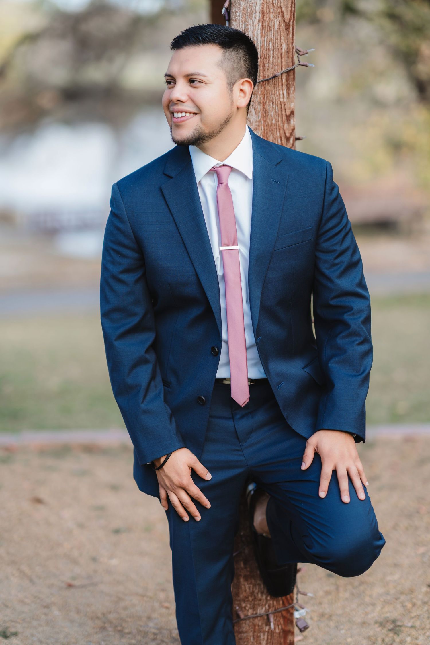 A person in a navy blue suit and pink tie poses playfully near a tree trunk in an outdoor setting.
